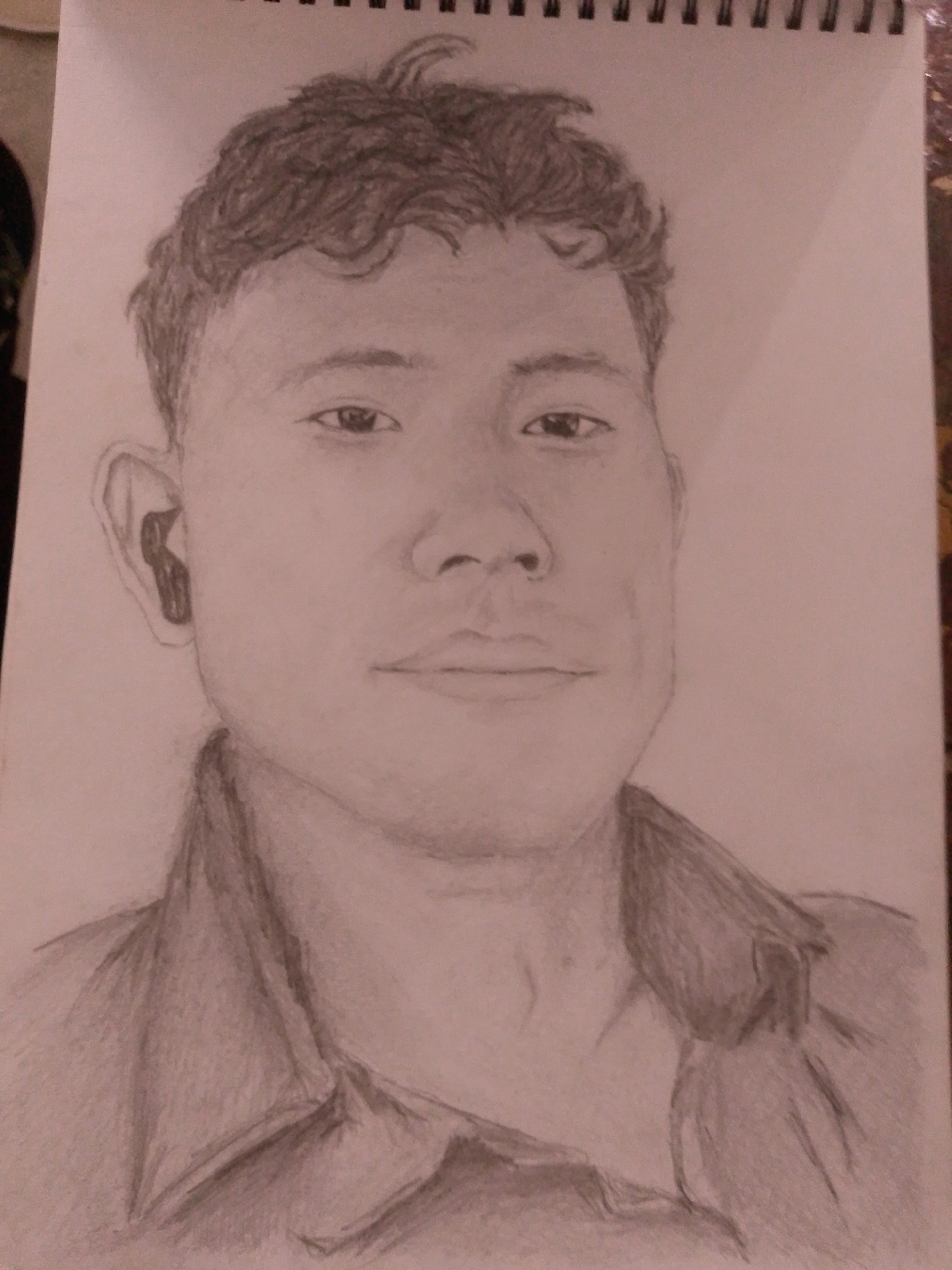 The image is a pencil sketch of a young man's head and shoulders. The drawing is done on a white sketchbook page. The man has dark, curly hair, dark eyes, and is wearing a collared shirt. He is also wearing an earbud in his left ear. The background of the sketch is blank. The sketchbook is spiral-bound at the top. There is no indication in the image that it is located in Sitarampuram, India.