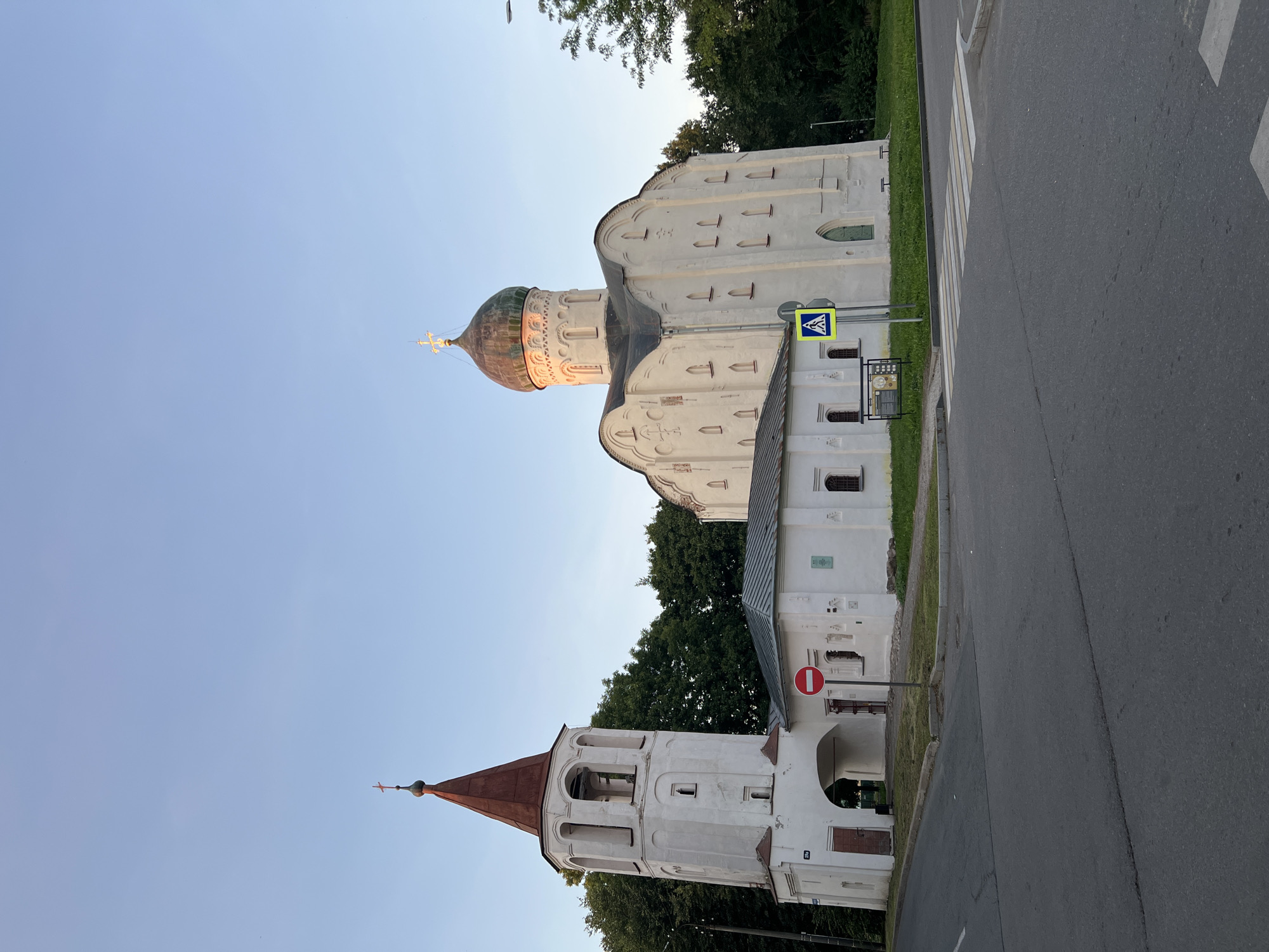 The image shows the Church of the Transfiguration of the Savior on Ilyina Street in Veliky Novgorod, Russia, under a clear blue sky. The church, constructed of white stone, features a prominent dome with a golden cross and a series of arched windows. Adjacent to the main church is a smaller, white building with a gray roof, and to the left, a white bell tower with a red roof and arched openings. A red and white circular "no entry" sign is visible on a pole in front of the smaller building. A pedestrian crossing is marked on the asphalt road in the foreground. Trees line the background, and the lighting suggests it is late afternoon or early evening, with the sun casting a warm glow on the side of the church dome. There are no people visible in the image.