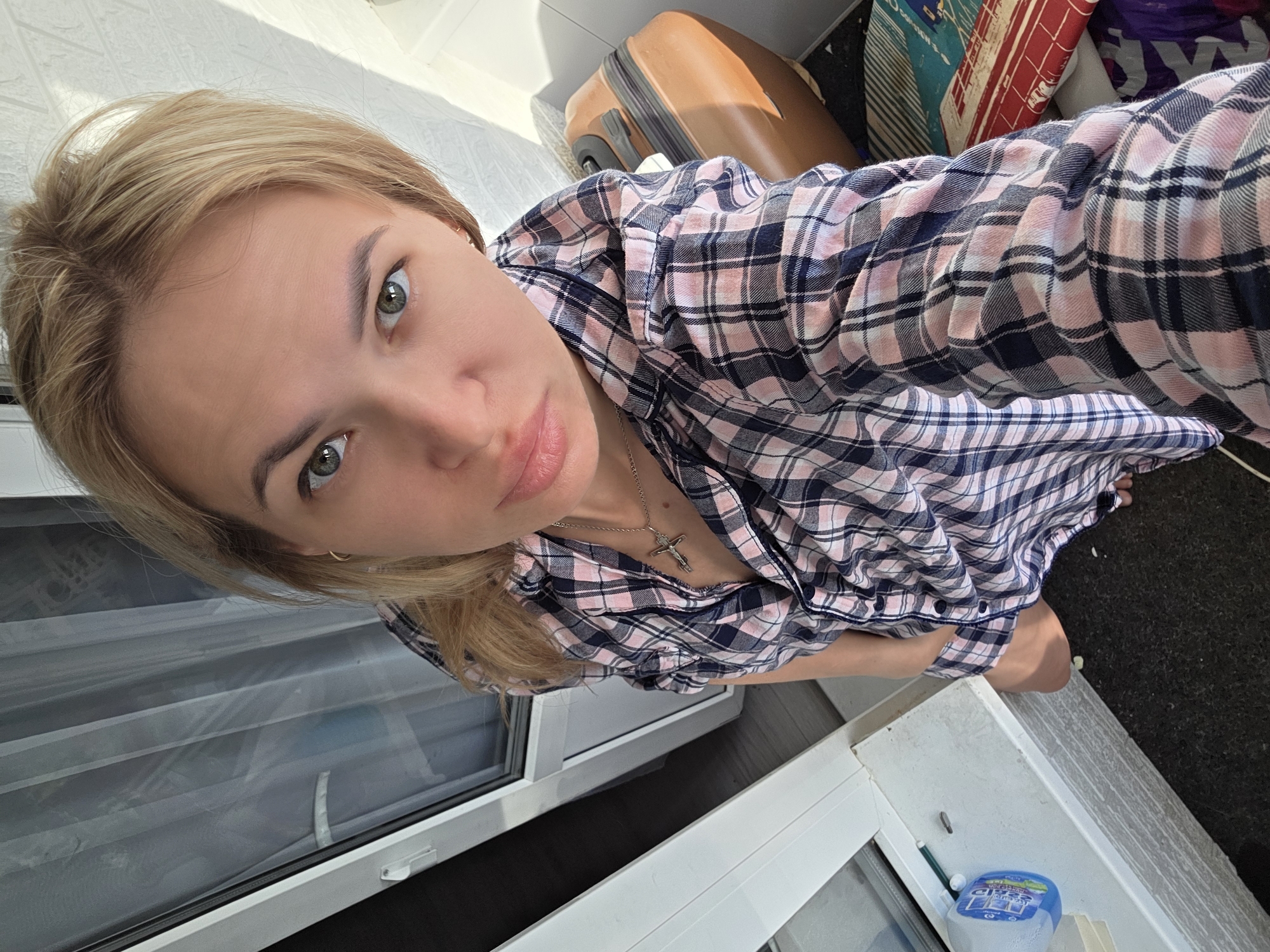 A young woman with blonde hair and green eyes is indoors in Chaikovsky, Russia. She is wearing a plaid shirt and a cross necklace. Behind her is a suitcase, a window with white frame and a bottle with blue lid.