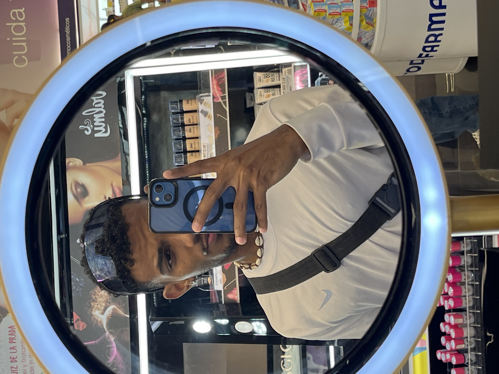 The image is a selfie taken in a mirror surrounded by a ring light, likely in a store setting, possibly a pharmacy, in Pampatar, Venezuela. A person with curly hair, wearing sunglasses, a white long-sleeved shirt with a Nike logo, and a shell necklace is holding up a blue smartphone to capture the reflection. The phone has a magnetic ring attached to the back. Behind the person and within the reflection, there are shelves stocked with various cosmetic and pharmaceutical products. Brands like "Cuida", "de la Prada", and "ODFARMA" are visible. Racks of lipsticks or similar products are on the right side of the reflection. The mirror seems to be part of a promotional display.