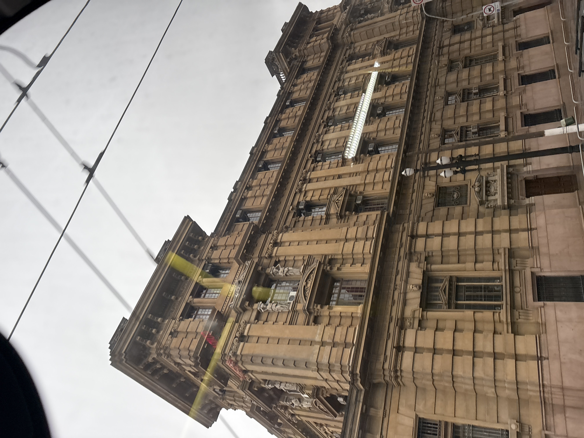 The image is an exterior, low-angle shot of a large, ornate, tan-colored building with multiple stories and visible architectural details. The building appears to be in the city of São Paulo, Brazil, based on its architectural style, which is reminiscent of historical public buildings in the city center.
Key architectural features include:
* **Facade:** The facade is characterized by a repeating pattern of pilasters, cornices, and recessed sections, creating a textured and layered appearance. The stone appears to be a light brown or tan color, possibly sandstone or a similar material.
* **Windows:** Numerous windows are visible, many with dark frames and some with visible air conditioning units installed. The windows vary in size and shape, with some being tall and narrow, while others are wider and more rectangular.
* **Statuary:** There are several visible statues integrated into the facade, particularly on the upper floors. These appear to be classical in style, depicting human figures.
* **Roofline:** The building has a prominent, stepped roofline with decorative elements and what appear to be small turrets or finials.
* **Entrance:** A large wooden door is visible at ground level, suggesting a main entrance.
The environment surrounding the building is partially visible:
* **Overhead Wires:** Numerous dark overhead wires, possibly for public transportation like trams or trolleybuses, cross the sky above the building. These are casting shadows on the building's facade and on the sky itself.
* **Street Furniture:** A dark lamppost with multiple lights is positioned in front of the building, and parts of railings or barriers are visible along the ground.
* **Signage:** A circular sign with a red border and what looks like a black graphic is visible on a pole on the right side of the image.
* **Sky:** The sky is overcast and bright white, indicating a cloudy or hazy day.
The image appears to have been taken from a moving vehicle, likely a bus or tram, due to the slight blur and the perspective. There are some light flares or reflections on the building, suggesting the photograph was taken through glass. No people are clearly discernible in the image.