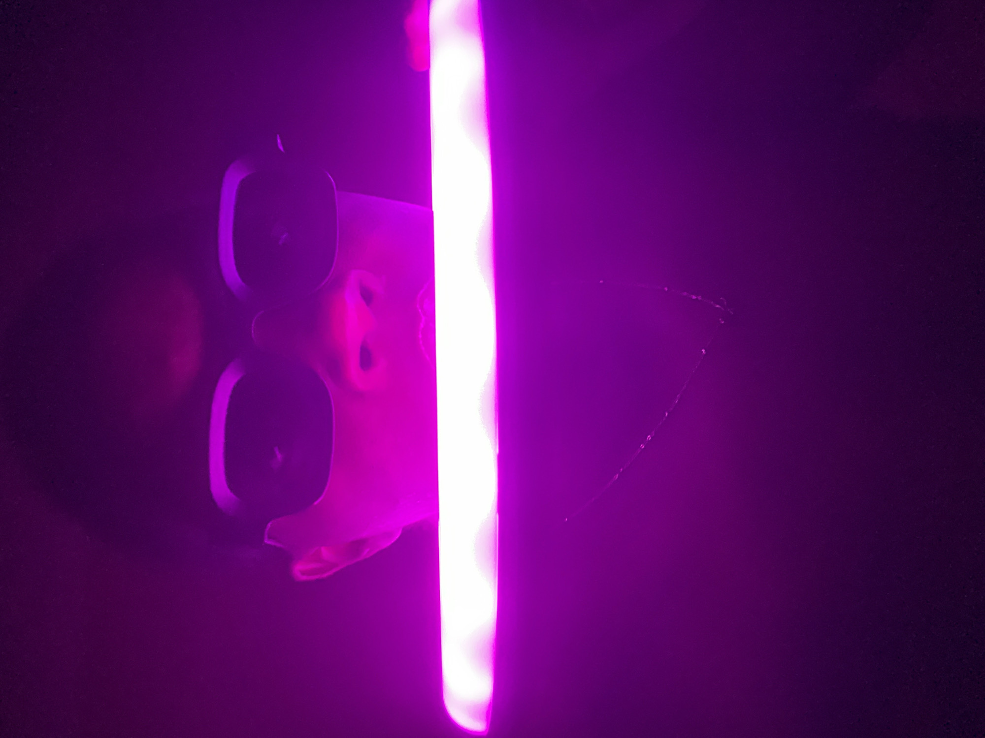 A person wearing rectangular glasses holds a bright, white LED light strip in front of their face in a dark environment. The image is dominated by hues of purple. The location is unknown.