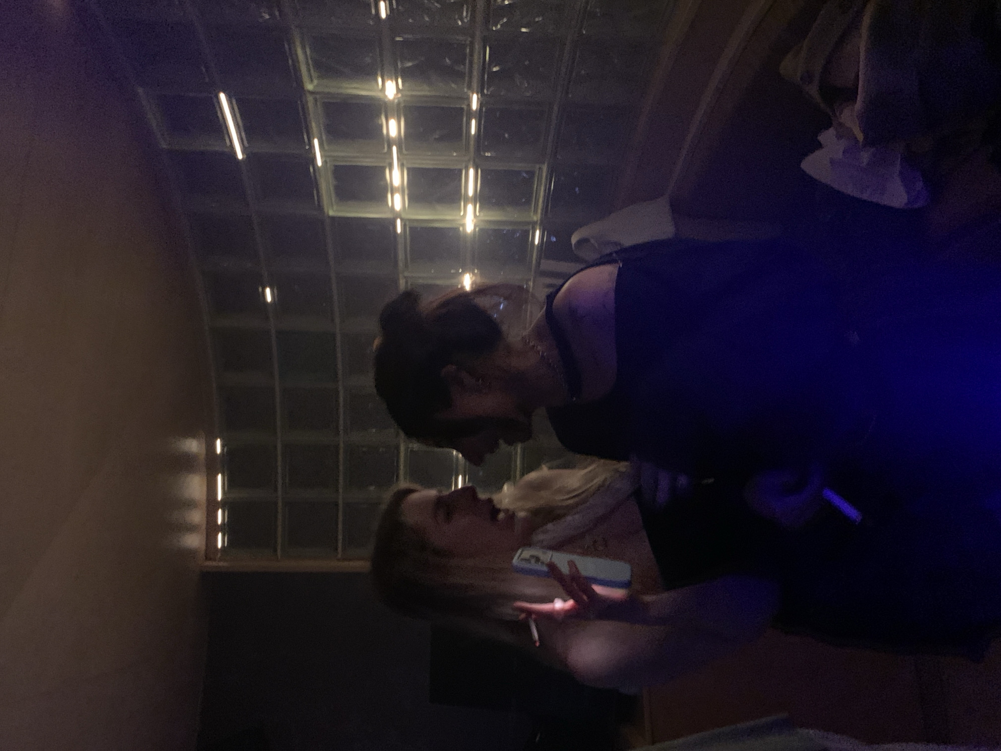 Two people stand close to each other indoors. One person has dark hair and is wearing a dark top, facing the other person, who has blonde hair and is holding a phone and a cigarette. The lighting is dim with a grid of lights visible in the background. No specific information about the location in Tel Aviv, Israel, is discernible.