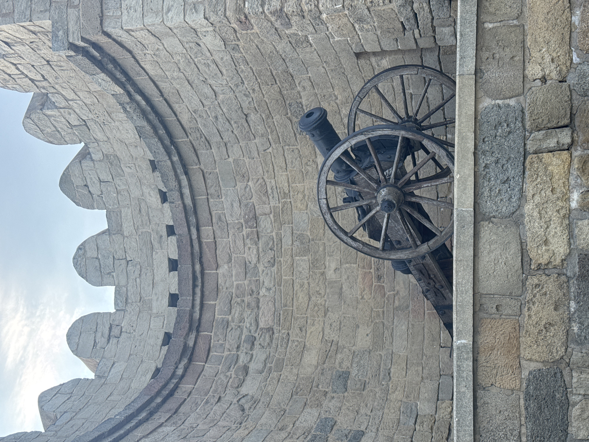 The image shows a historical stone cannon mounted on a stone wall. The cannon is old, with wooden wheels and a metal barrel. It is positioned in an alcove within a stone structure, likely a fortress or defensive wall. The stone architecture is characterized by rough-hewn blocks and crenellations at the top. The background is a cloudy sky. This cannon is located in the Sabail Raion of Baku, Azerbaijan, as part of the historical fortifications. There are no people visible in the image, and the scene is static.