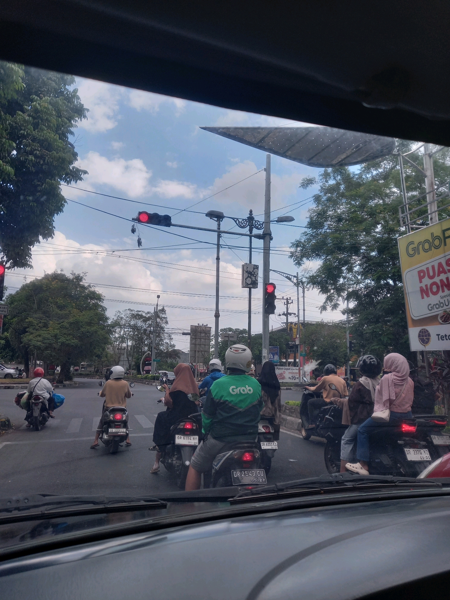 This image shows a street scene in Mataram, Indonesia, likely at an intersection, with several motorcycles and their riders. The traffic lights are red, indicating that traffic is stopped. The sky is partly cloudy.
In the foreground, the windshield of a car is visible, with the dashboard below it. The perspective is from inside the car, looking forward.
Several motorcycles are stopped in front of the car. On the left, a rider in a white helmet is on a motorcycle carrying luggage. In the center, a rider in a tan shirt and white helmet is on a motorcycle. To the right, a rider in a brown hijab is on a motorcycle, with a passenger behind her wearing a pink hijab. Further right, a rider in a green Grab jacket is on a motorcycle.
In the background, there are trees, buildings, and streetlights. A large yellow sign for "GrabFood" is visible on the right. There are also several power lines and traffic signal poles. The road surface is asphalt, and a pedestrian crossing is marked on the road. License plates are visible on some of the motorcycles, including "DR 3208 E1," "DR 6194 NH," "DR 2543 CU," and "DR 6727 ZU."