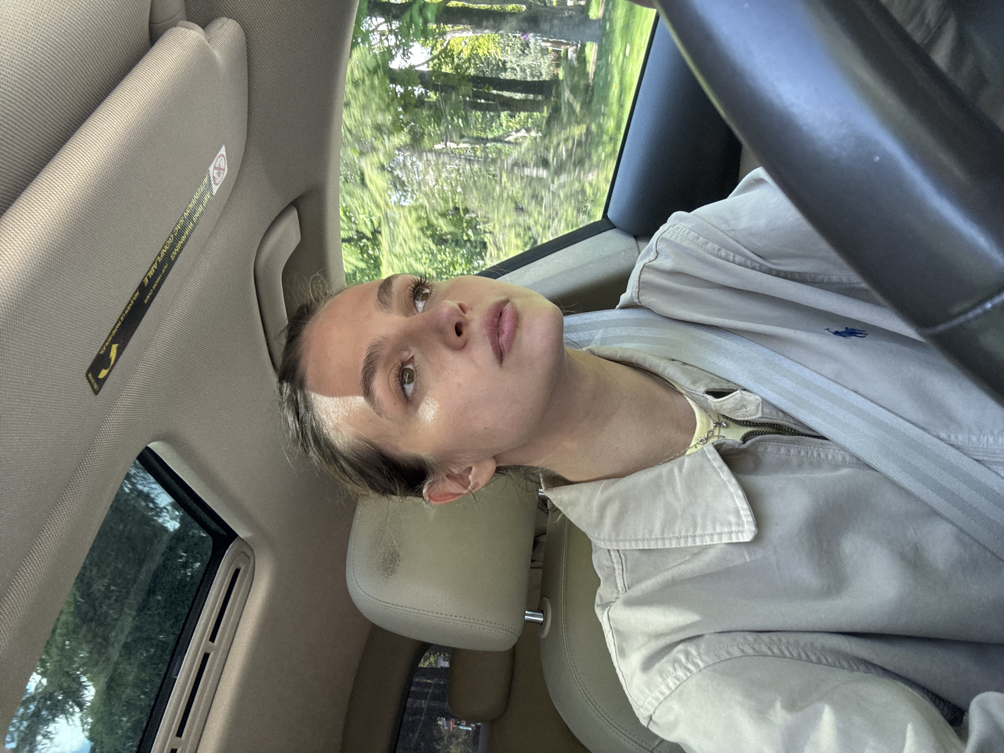 A woman is in the driver's seat of a car. She is wearing a light-colored jacket with a Polo Ralph Lauren logo visible. The car is moving, as suggested by the blurred background visible through the window. The location is Unknown, Unknown.