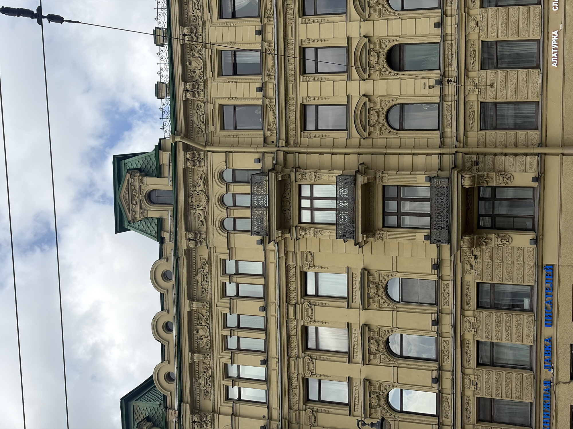 The image depicts a portion of a building facade in Saint Petersburg, Russia. The building is light yellow and adorned with intricate architectural details, including decorative moldings and ornate window surrounds. There are multiple windows visible, some with balconies. The sky is visible in the background, with a mix of clouds and patches of blue. Utility wires run diagonally across the top left of the frame. A sign with text in Cyrillic is on the building, which translates to "Alatyrka". The angle of the photograph gives an upward perspective of the building. The overall environment appears urban.