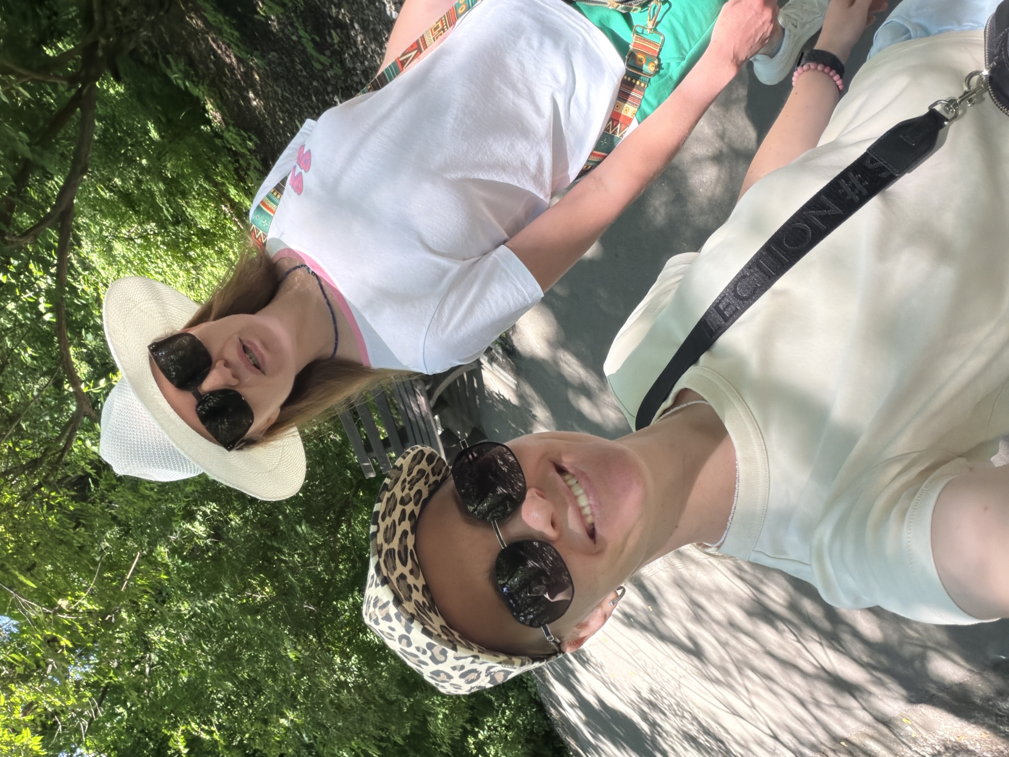 Two women are posing for a selfie outdoors. The woman in the foreground, wearing a leopard-print cap and sunglasses, smiles at the camera. She has on a light-colored t-shirt and a black crossbody bag with a patterned strap. The woman behind her, wearing a white sun hat and sunglasses, is looking to the side. She has on a white t-shirt with colorful trim and green pants. They appear to be in a park or wooded area with dappled sunlight on the ground. There is a wooden bench visible in the background. The location is not specified as Khabarovsk, Russia, nor is there any information to suggest this.