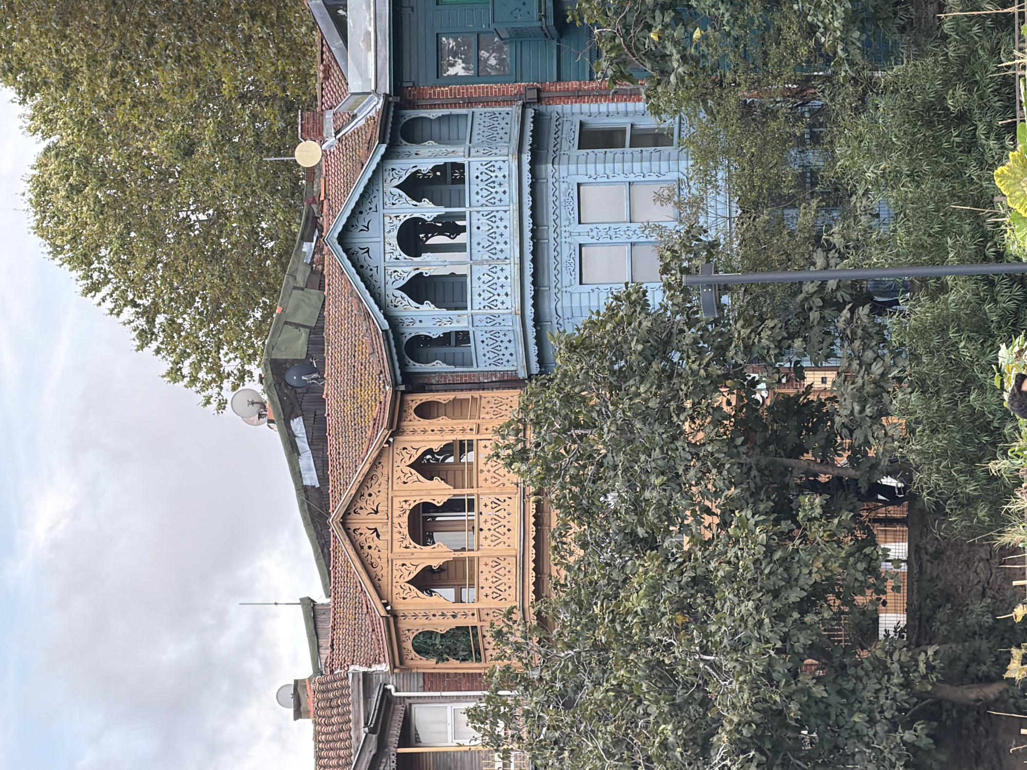The image shows a close-up view of two ornately decorated wooden houses in Üsküdar, Turkey, with lush green trees in the foreground. The house on the left is painted a warm, light brown, and features intricate wooden latticework on its bay window, which is adorned with arched openings and decorative patterns. The house on the right is painted a vibrant, sky blue and has a similar bay window with detailed carvings. Both houses have tiled roofs and are situated close together, suggesting they are part of a row of houses. Behind these structures, larger trees with dense foliage can be seen against a cloudy sky. The overall scene depicts traditional architecture nestled within a verdant environment.