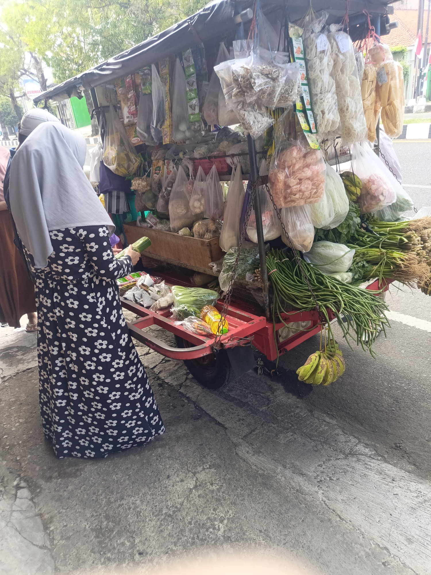 An outdoor street vendor stall is shown in Desa Gunungsimping, Indonesia, featuring a variety of fresh produce and packaged goods. The stall is on wheels and has a dark tarp for cover. A woman wearing a hijab and a floral print dress is inspecting a cucumber at the stall. The ground is paved with concrete. Various vegetables such as long beans, cabbage, and leafy greens are visible, as are bananas hanging from the cart. Packaged snacks and dried goods are displayed on hooks above the produce. The scene suggests a typical market day in a local Indonesian community.