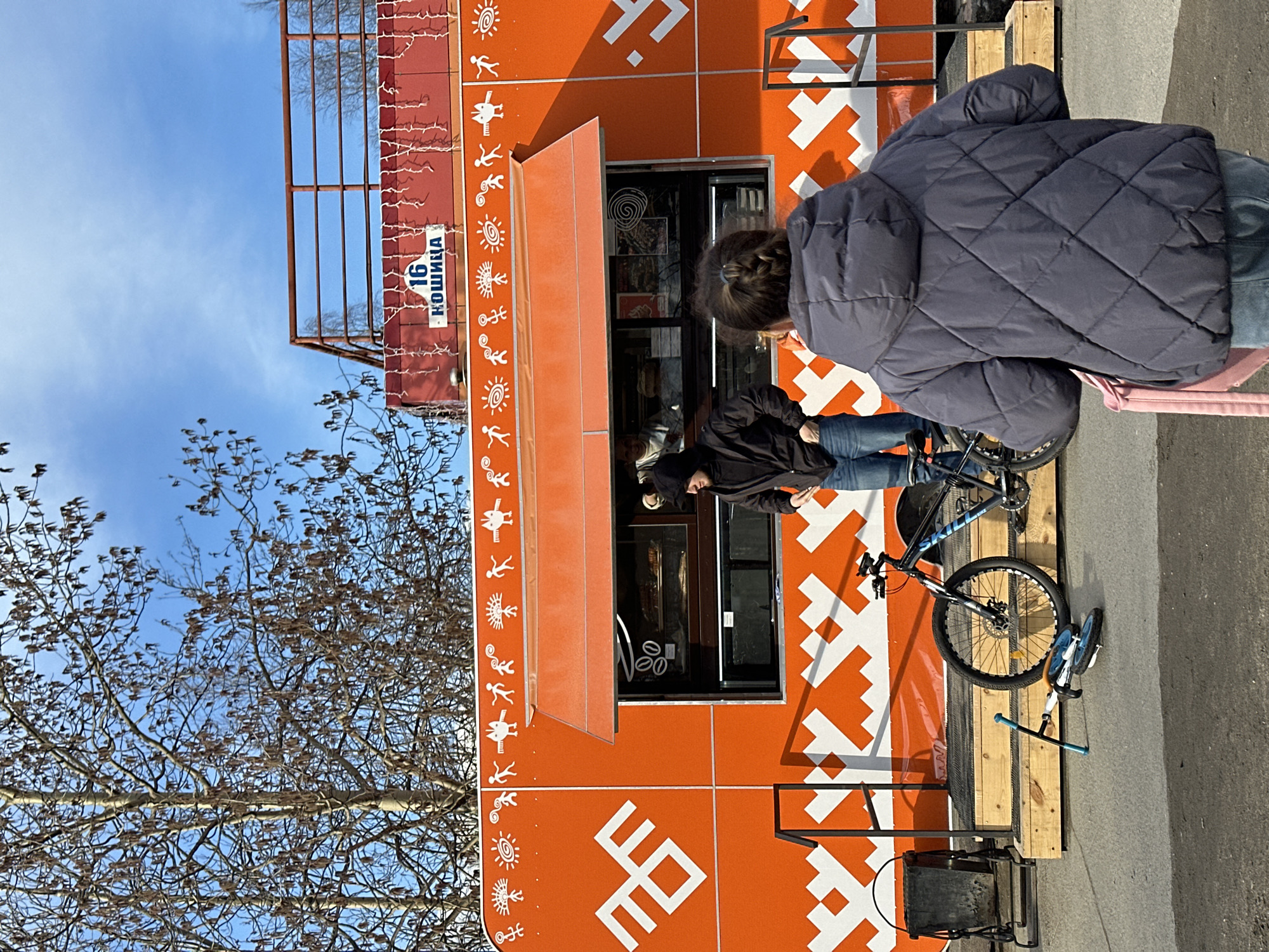 The image shows a street scene in an unknown city. An orange food kiosk with white decorative patterns is situated next to a sidewalk. There is a sign on the kiosk structure with a street address that appears to be "16 Кошица". Two people are standing at the serving window of the kiosk, seemingly engaged in a transaction. One is wearing a black jacket and jeans, and the other is wearing a dark quilted jacket and jeans. A black bicycle with blue accents is parked next to the kiosk. To the left, there is a tree with bare branches against a blue sky. A metal structure is attached to the top of the kiosk. Part of the street and sidewalk are visible in the foreground.