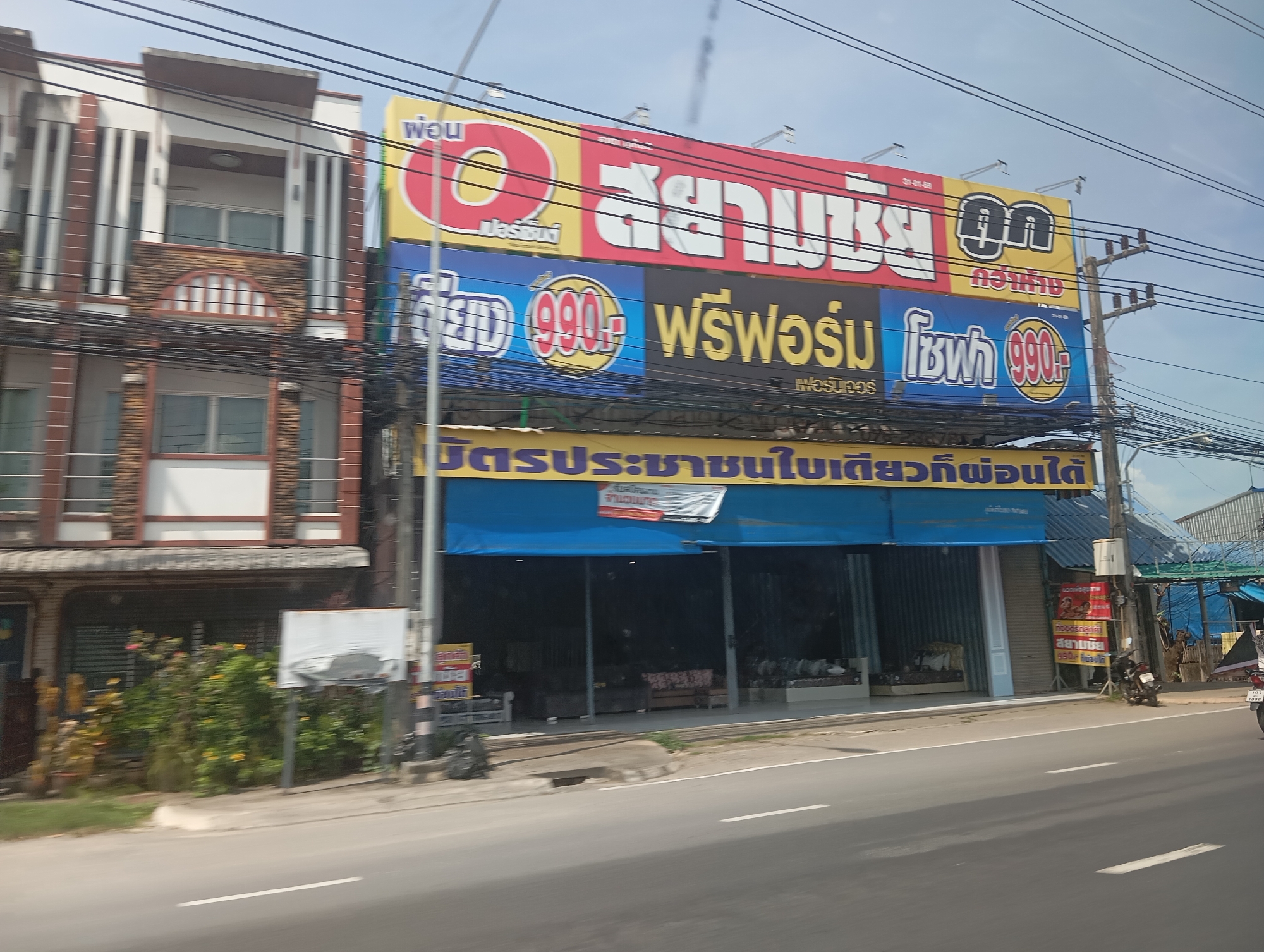 In the city of Ko Kaeo, Thailand, a building with multiple levels stands beside a road. The building features a white facade with brown accents, and its upper floors have balconies and windows. A large, colorful sign is prominently displayed on the building's exterior, featuring Thai text and advertisements for mattresses and furniture, including images of mattresses and the price "990". Below the main sign, a blue awning covers the entrance to the establishment, and inside, several mattresses are visible. A power pole with numerous wires runs alongside the road. To the right of the entrance, a smaller red sign advertises a product with Thai text and images. The scene is captured from a moving vehicle, as indicated by the motion blur.