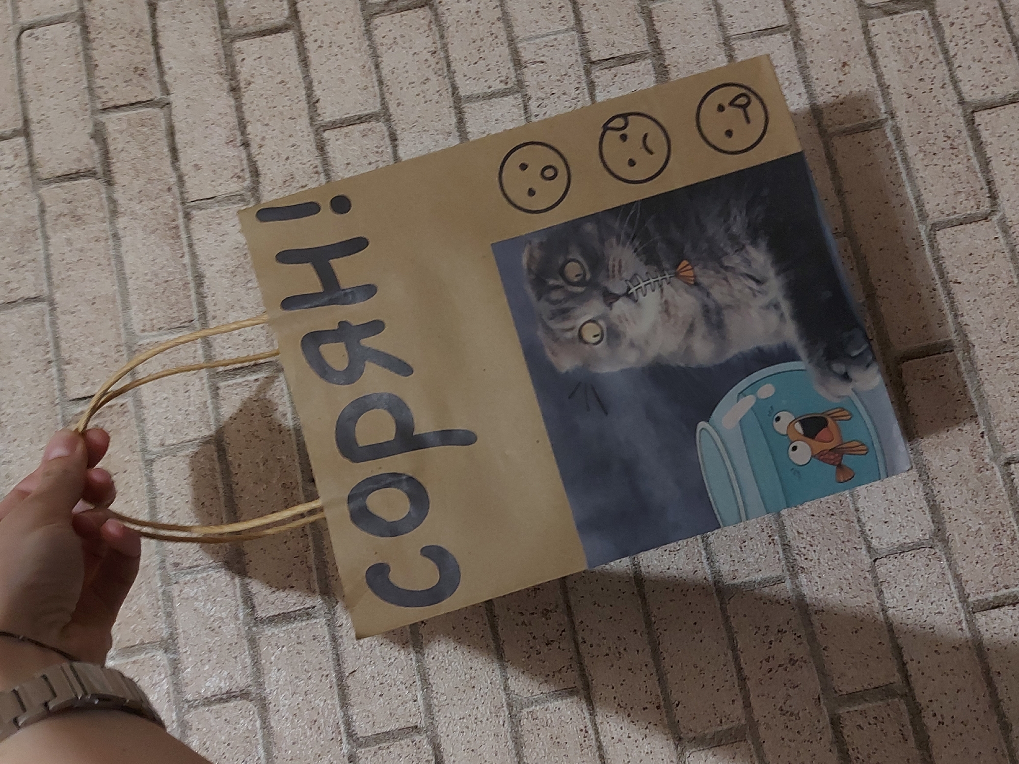 A brown paper bag with drawings and text is being held by a person. The bag has three cartoon faces drawn on the top, and a picture of a cat looking into a fishbowl is printed on the front. The Russian word "сорян!" (soryan!, sorry!) is written vertically in black marker on the side of the bag. The person holding the bag is wearing a watch on their wrist. The background is a textured brick surface. Based on previous information, the location is Izhevsk, Russia.