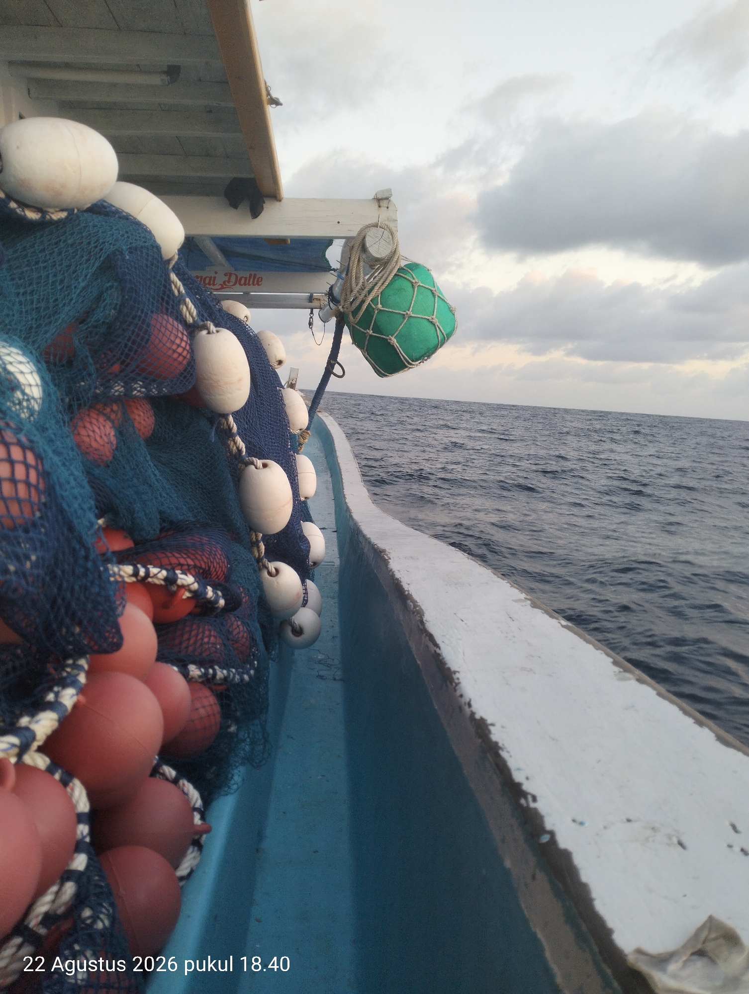 The image shows the side of a fishing boat on the ocean. The boat is blue and white. On the deck of the boat, there are blue fishing nets and many buoys. The buoys are white and red. There is also a green buoy hanging from the side of the boat. The ocean is dark blue and there are some clouds in the sky. The photo was taken on August 22, 2026, at 6:40 PM.