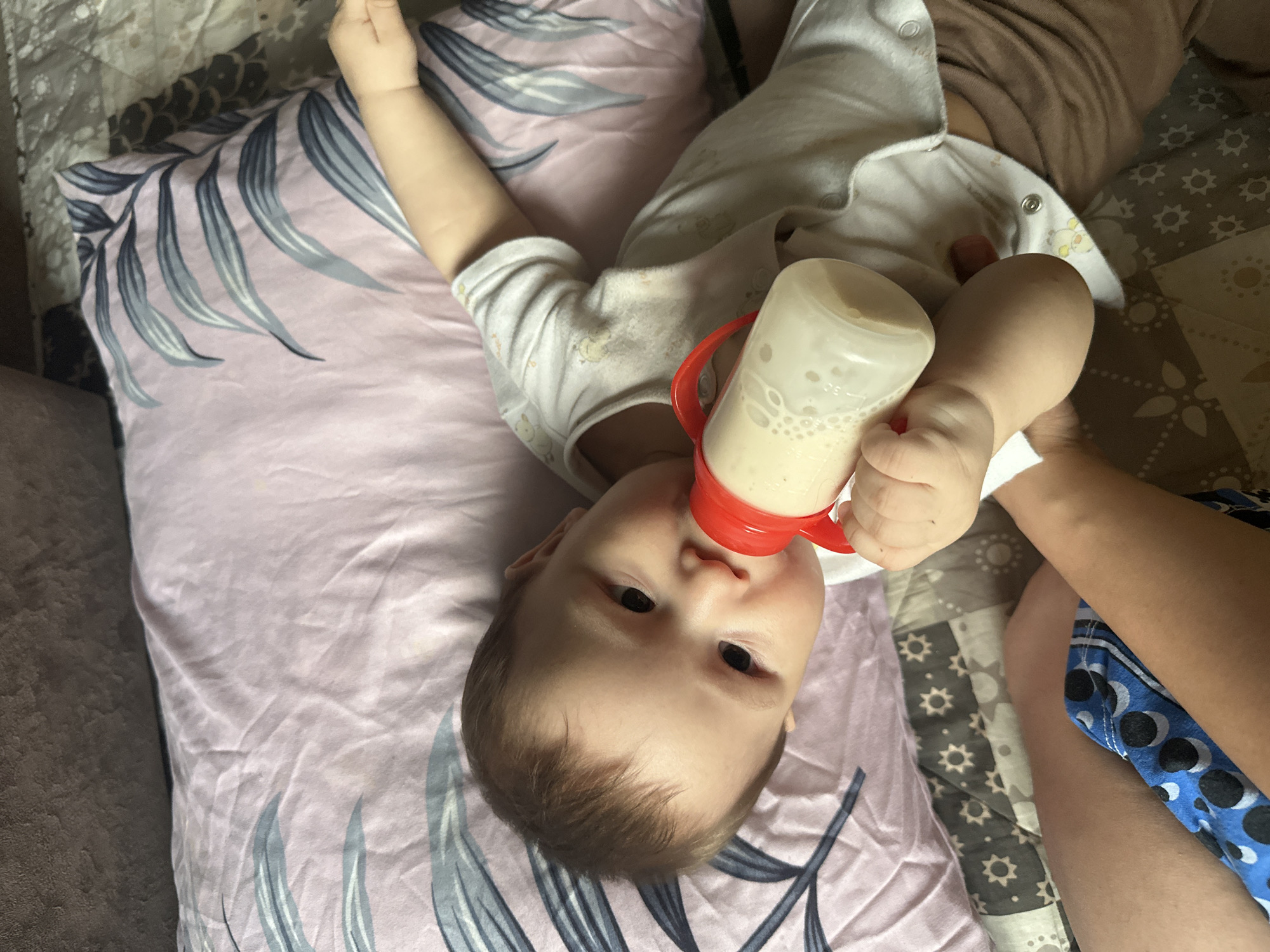 An overhead shot captures a baby lying on their back, drinking from a white bottle with a red handle. The baby, appearing to be an infant or young toddler, is positioned with their head towards the bottom of the frame and their body extending upwards. They wear a white long-sleeved onesie with small, light-colored animal patterns and snaps down the front, paired with light brown pants. Their dark eyes look directly towards the camera with a neutral, calm expression.
An adult's hand and arm are visible on the right side of the frame, holding the bottle steady for the baby. The bottle contains a light cream-colored liquid, likely milk or formula, with visible bubbles. Volume markings "30", "60", and "90" are faintly visible on the side of the bottle.
The baby is resting on a light pink pillow decorated with a grey and blue leaf pattern. To the right of the baby, a grey blanket or sheet with a repeating pattern of small stars or sunbursts is visible. Part of a blue fabric with a dark blue and black circular pattern, possibly clothing or another blanket, can be seen on the far right. The setting appears to be indoors, likely a home, with soft, diffused lighting.