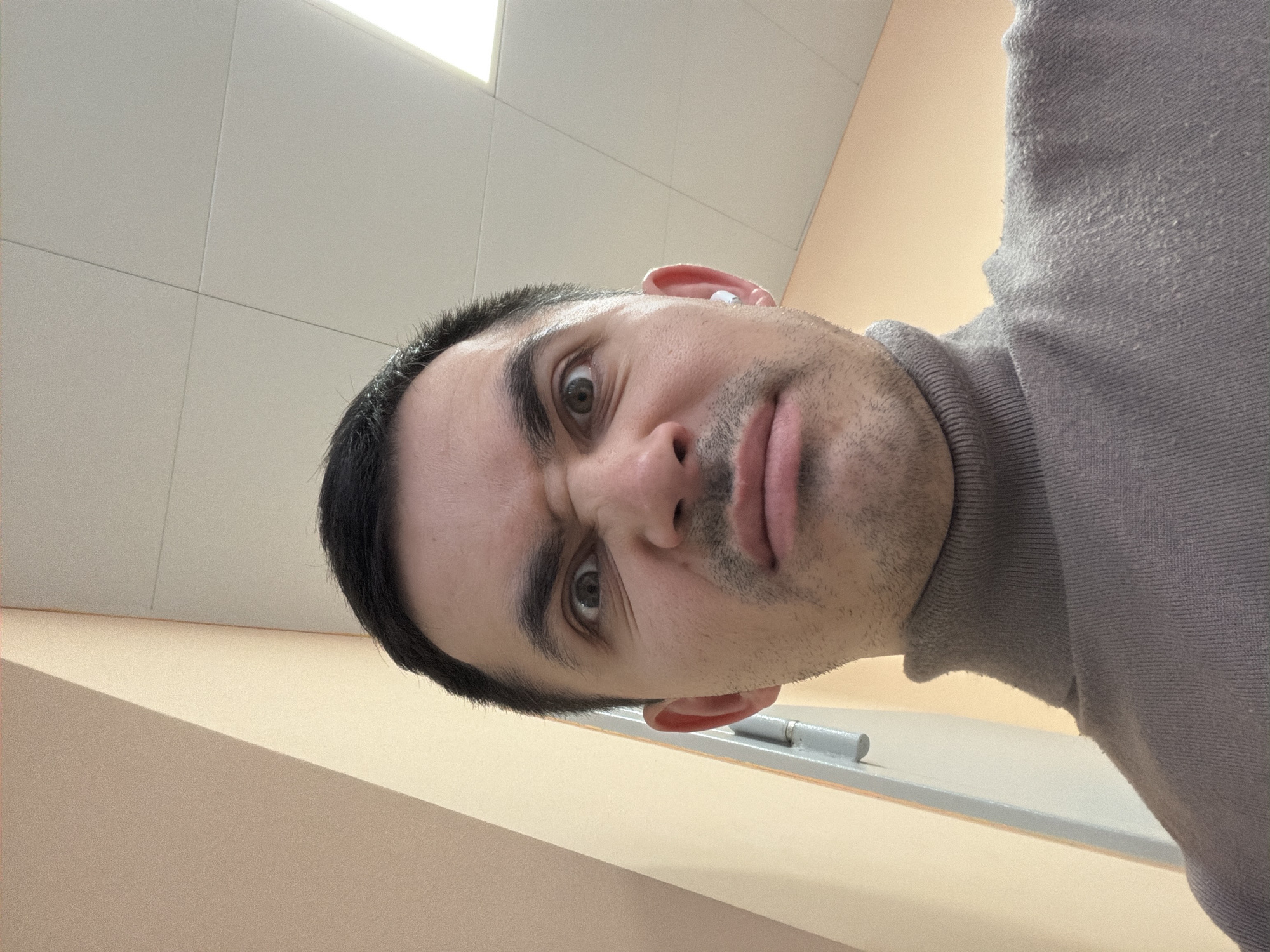 A close-up, low-angle selfie of a man with dark hair, light skin, and brown eyes. He is wearing a gray turtleneck sweater. His face is slightly contorted into a questioning or skeptical expression, with furrowed brows and a pursed mouth. He appears to be indoors, with a white tiled ceiling and a beige wall visible in the background. A gray object is visible near his face and ear. The specific location in Izhevsk, Russia, cannot be determined from the image.