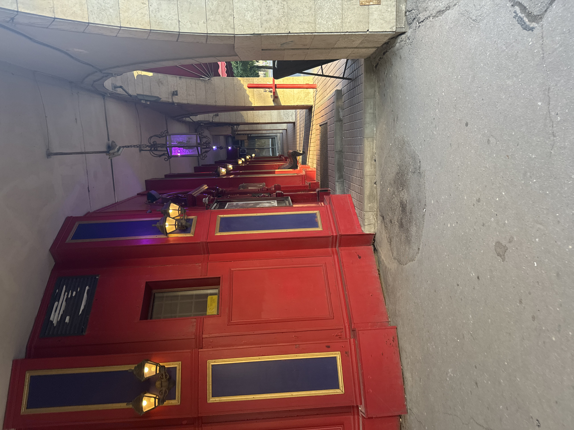 The image shows an outdoor walkway in front of a red building with ornate gold trim and blue panels. Several vintage-style lanterns are mounted on the red wall, casting a warm glow. Further down the walkway, a decorative purple lantern hangs from the covered ceiling. The walkway is paved with brick and is bordered by arched stone columns on the right. Sunlight casts long shadows across the walkway. A dark easel is positioned near the entrance to the walkway on the right. The red building appears to be a commercial establishment, possibly a bar or restaurant, given the decorative facade and lighting. The environment suggests an urban setting, likely in a historic district given the architectural style. No people are visible in the image.