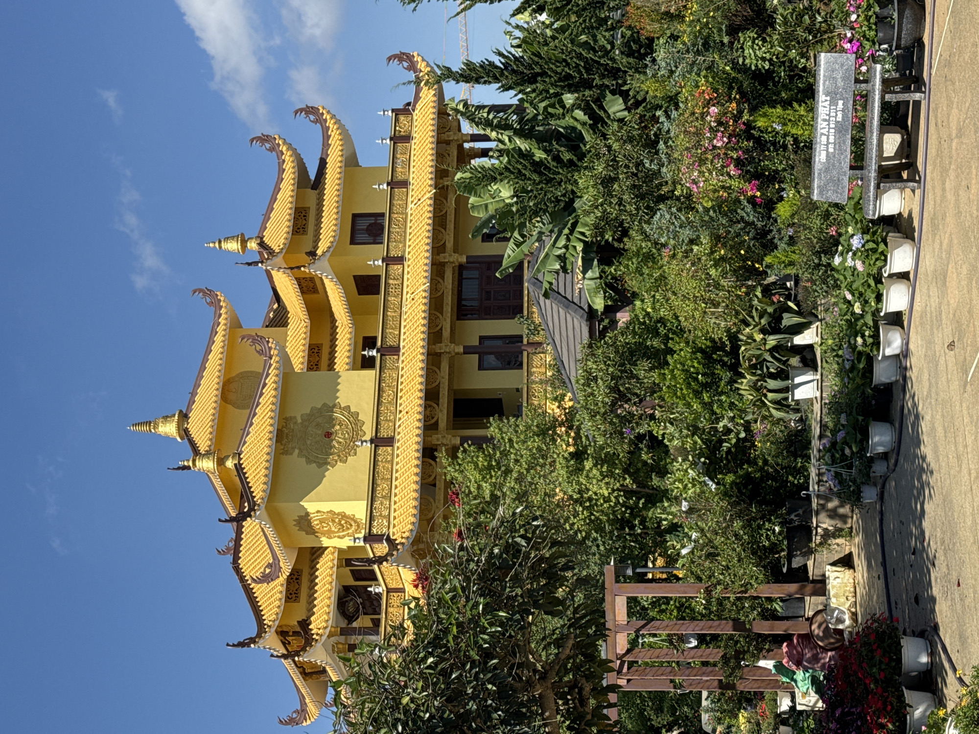 This image captures a vibrant, multi-tiered golden building, likely a pagoda or temple, under a clear blue sky in Da Lat, Vietnam. The building's architecture features elaborate golden ornamentation, including ornate curved eaves, intricate patterns on its yellow walls, and decorative finials topping its multiple roof levels. Dark wooden window frames and doors punctuate the building's facade.
The structure is nestled within a lush garden brimming with diverse flora. Dense green foliage, including tall trees, bushes, and flowering plants, envelops the building. Various potted plants, displaying colorful blooms in shades of pink, purple, and red, are arranged on a paved surface to the right. Some larger plants resembling banana trees are also visible.
No specific human activity is depicted; the scene presents a static view of the architectural site and its surrounding landscape. The time of day is bright daylight, indicated by the clear sky with a few wispy clouds and the distinct shadows cast on the ground. A faint outline of a construction crane is visible in the distance, partially obscured by the trees.
A rectangular gray stone slab or sign stands among the plants on the right. Visible text on this slab includes Vietnamese words: "CÔNG TY TNHH AN PHÁT" (AN PHAT CO., LTD), a phone number "DTC: 0918 013 811", "ĐÀ LẠT", and the enigmatic phrase "NOI 1 TON".