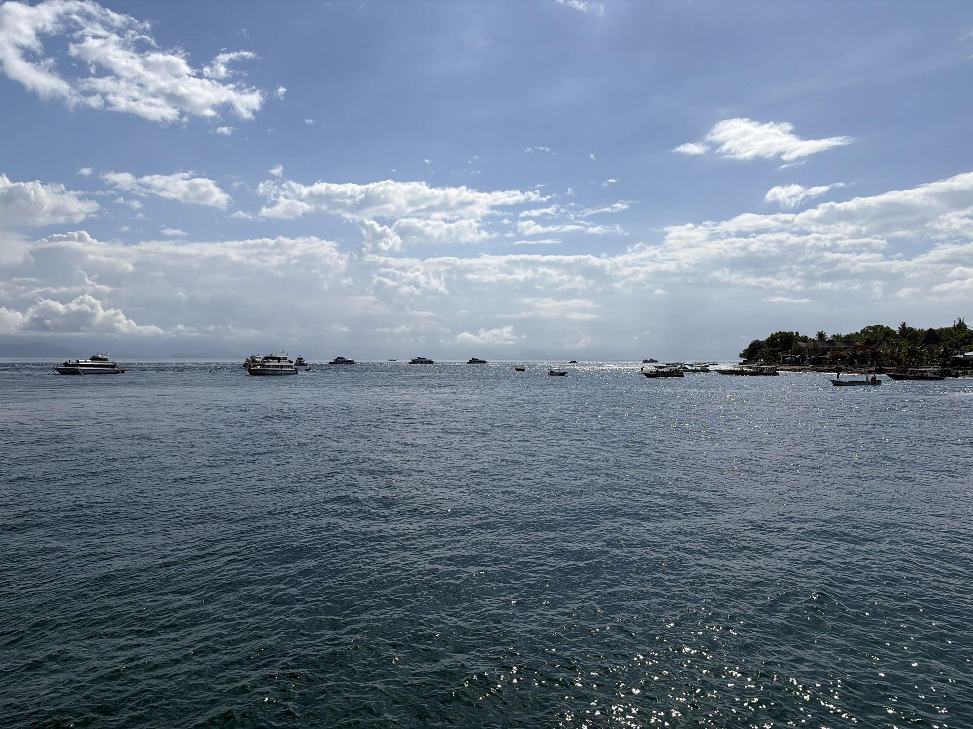 The image captures a wide-angle view of a body of water, likely the ocean, with several boats of various sizes dotting the surface. In the distance, a lush green coastline is visible, dotted with buildings and palm trees, suggesting a tropical location. The sky is a vibrant blue, partially filled with scattered white clouds. The water itself is a deep blue, with sunlight glinting off its surface, creating a shimmering effect. The scene appears peaceful and serene, likely depicting a coastal area or harbor. The location is identified as Kampung Toyapakeh, Indonesia, known for its beautiful beaches and waters.
