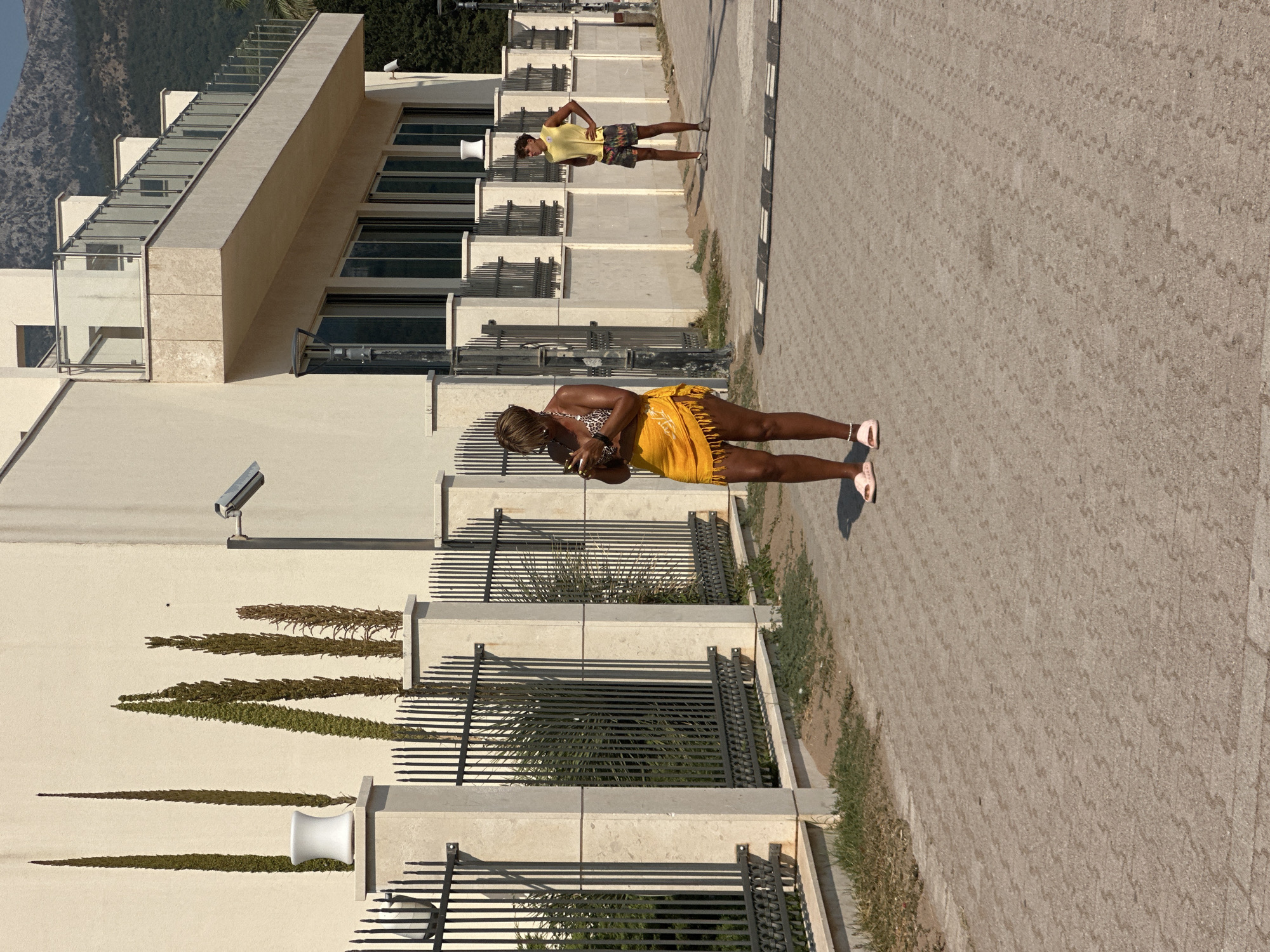 In Boreti, Montenegro, on a sunny day, a woman stands on a paved walkway in front of a modern building with a metal fence. She is wearing a leopard-print bikini top, a yellow sarong skirt with fringe, and pink slides. She is looking down at her phone, which she holds in her hands. Behind her, the building features a balcony with a glass railing and a security camera is visible on a pole attached to the building. Further in the background, another person, a young man wearing a yellow sleeveless shirt and patterned shorts, stands with hands on his hips. The environment includes tall, slender trees planted in front of the building and a mountainous landscape in the distance.