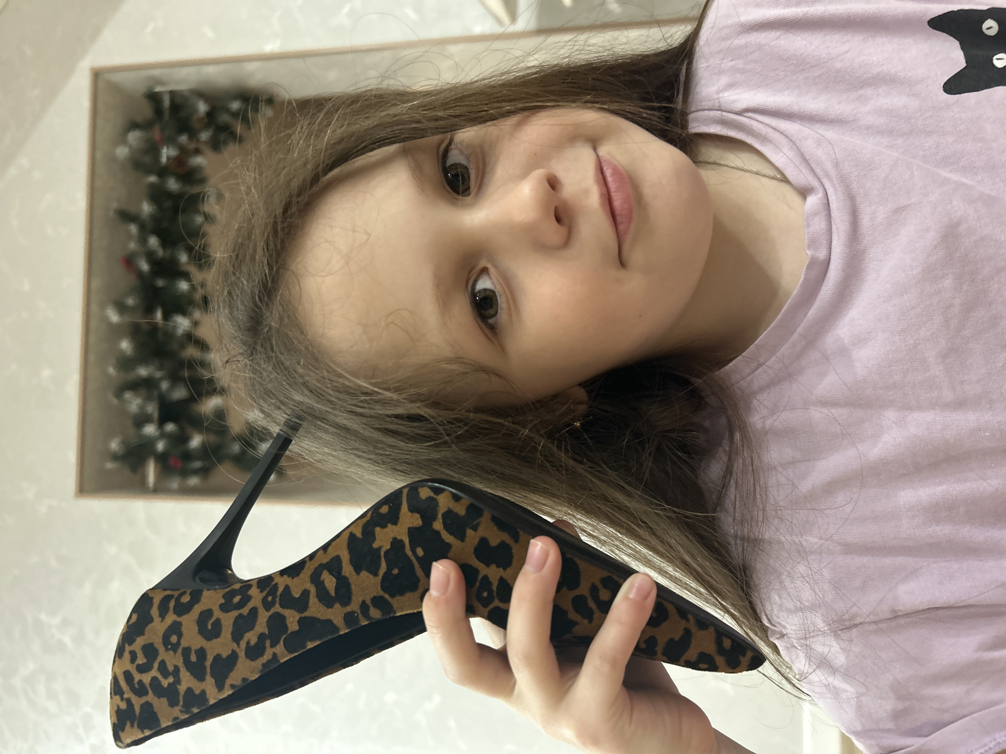 A young girl is depicted in an indoor setting, playfully holding up a high-heeled shoe. The image is oriented with the girl's head on the left side, looking directly at the camera with a gentle smile. She has long, straight brown hair, light skin, and brown eyes. She is wearing a light lavender or purple t-shirt, which features a partial black cat design on the upper right side.
In her right hand, the girl is holding a stylish high-heeled pump shoe. The shoe features a distinct leopard print pattern in shades of brown and black, and has a sleek black stiletto heel. Her fingers are visible gripping the side of the shoe.
The background is a bit blurry but reveals elements suggesting an indoor environment. Behind the girl's head and the shoe, a reflection in what appears to be a framed mirror or picture on a wall shows part of a Christmas tree or festive green garland with red accents, indicating that the photo might have been taken during the holiday season. The lighting is soft, consistent with indoor conditions. The overall mood conveyed is one of youthful playfulness. This scene takes place in Chaikovsky, Russia. There is no visible text in the image.