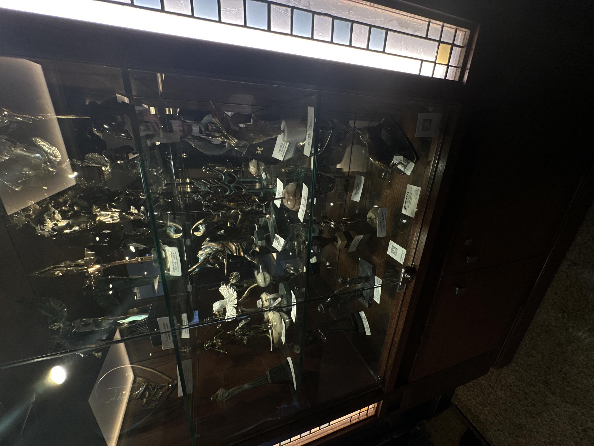 A dimly lit, full, slightly angled shot depicts a glass display case situated in an unspecified building in Sofia, Bulgaria. The cabinet is filled with a collection of items, potentially artifacts or sculptures, made of a reflective, metallic material. White labels or tags are affixed to some objects. The background behind the case is dark, making it difficult to discern details beyond the cabinet. Above the case is a window with translucent tiles. The right side of the image shows the wooden side of the display case, which is adjacent to a dark wall. The floor below the cabinet appears to be carpeted.
