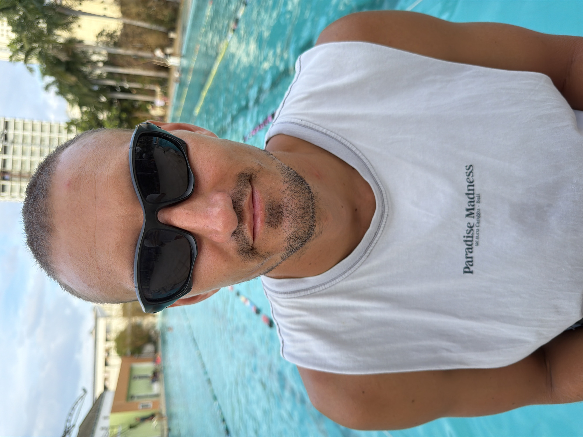 A man wearing sunglasses and a white tank top stands in front of a swimming pool. The words "Paradise Madness, SE ALCO Canggu - Bali" are printed vertically on his shirt. In the background, there are buildings and palm trees. The location is Nha Trang Ward, Vietnam.