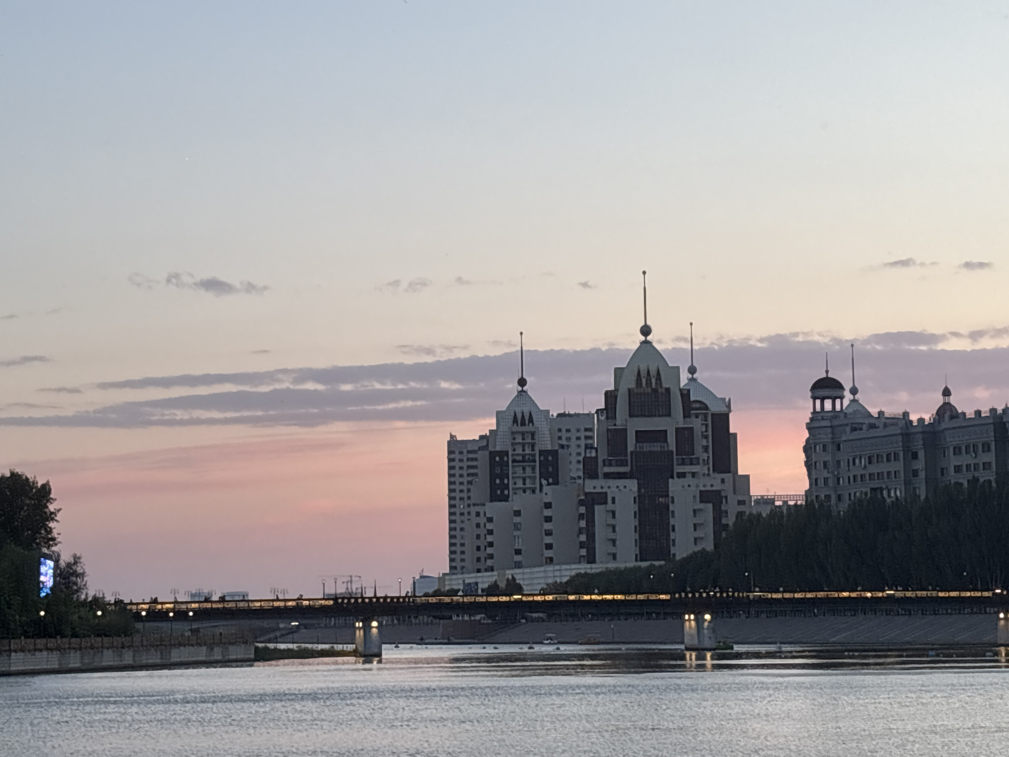 This is a wide-angle, eye-level photograph of a river in Astana, Kazakhstan, at dusk. The sky is a gradient of pale blue at the top fading to pink and then a soft orange near the horizon, with scattered, wispy clouds. The water in the foreground reflects the sky and the lights from the city. Across the river, a bridge spans the water, illuminated by a series of lights. Beyond the bridge, a collection of modern buildings rises above a line of trees. The buildings have varied architectural styles, with some featuring spires and distinct geometric designs. One prominent building is a multi-story structure with a central tower topped by a decorative spire, and it is flanked by other tall buildings. The overall scene conveys a sense of urban tranquility at the end of the day. There are no visible people in the frame.
