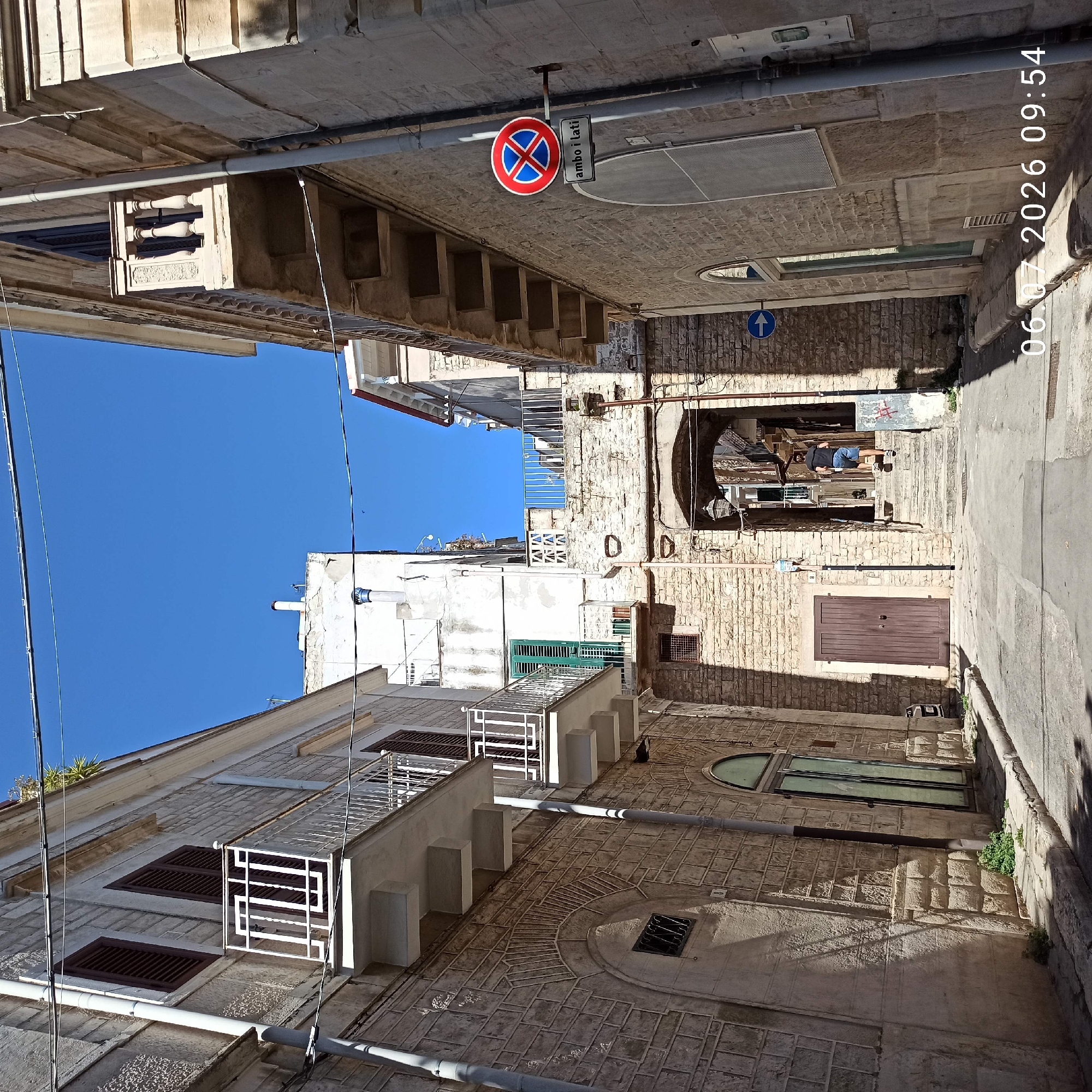 The image depicts a narrow street in Ruvo di Puglia, Italy, with stone buildings lining both sides. The street is paved and slopes gently upwards towards an arched passageway. On the left, a building features a stone facade, a wooden door, and a balcony with a white railing. On the right, another building also has a stone facade, a balcony with a decorative railing, and a circular "no parking" sign with Italian text below. A person in a black t-shirt and blue shorts is ascending a set of stone stairs through the arched passageway. The sky is clear and blue. The timestamp "06.07.2026 09:54" is overlaid on the bottom right of the image.