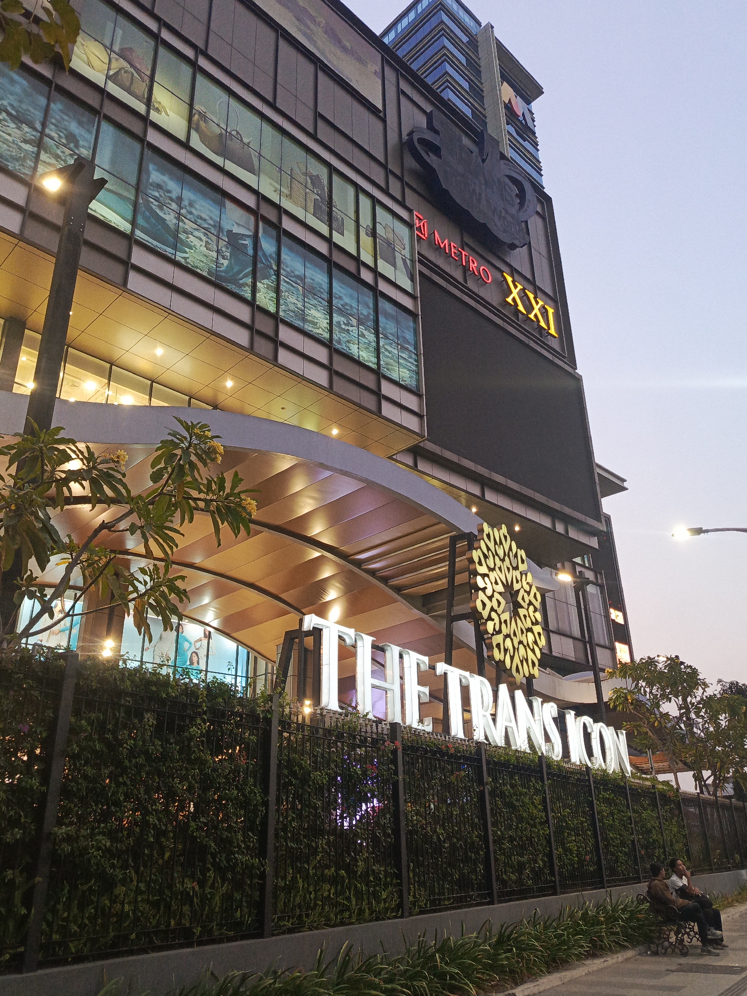 The image depicts the exterior of The Trans Icon building in Surabaya, Indonesia, likely at dusk. The building is a modern, multi-story structure with a dark facade. Several levels of windows are visible, some displaying images and others reflecting the sky. A large black logo, possibly representing a musical instrument, is mounted on the upper section of the building, above a red and yellow illuminated sign that reads "METRO XXI". Below this, a large circular golden emblem is prominently displayed. The words "THE TRANS ICON" are illuminated in white letters across the front of the building at ground level. A black metal fence, covered with green foliage, runs along the front of the property. Two individuals are seated on a bench in front of the fence, facing towards the building. The sky is a pale grey, and a street lamp is illuminated on the right side of the frame. Lush green plants and a tree with yellow flowers are visible in the foreground.