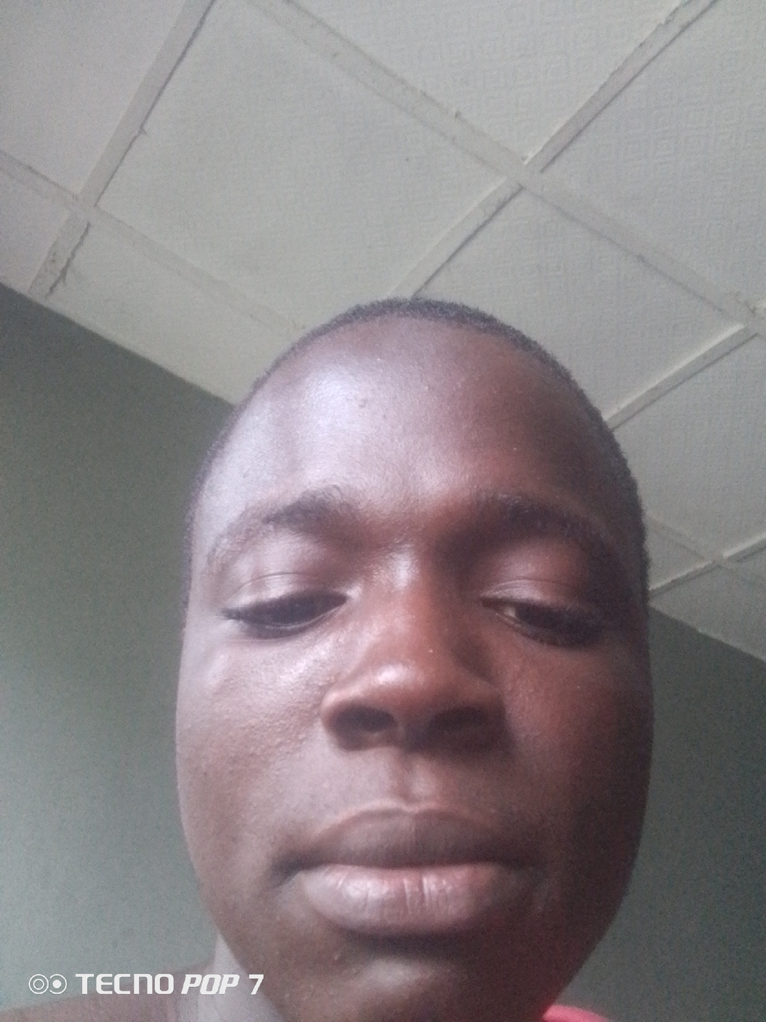 The image is a close-up, low-angle selfie of a young person's face, taken indoors. The background features a textured, off-white ceiling with a grid pattern. The walls appear to be a muted green color. The person has dark skin and short, dark hair. Their eyes are partially closed, and their mouth is slightly open, revealing their lower teeth. The lighting is somewhat dim, creating subtle shadows on the face. In the lower-left corner, there is a watermark that reads "TECNO POP 7". The image quality is grainy, suggesting it was taken with a mobile phone camera in low light conditions. The context of Ipetumodu, Nigeria, implies this is likely a domestic indoor setting.