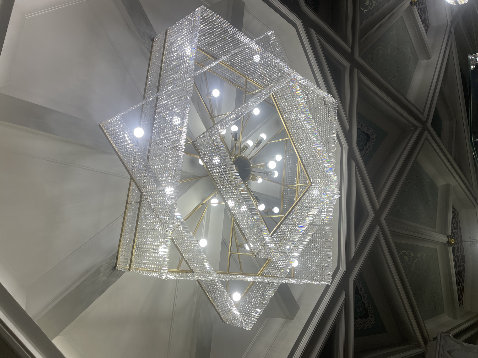 A low-angle shot captures a grand, ornate chandelier in Kazan, Russia. The chandelier is multi-tiered, featuring stacked, angular frames constructed from gold-colored metal. Each tier is draped with strings of clear crystals, creating a shimmering effect. The chandelier's center features multiple light bulbs. The ceiling, visible in the background, is decorated with geometric patterns, some sections painted with ornate floral designs.