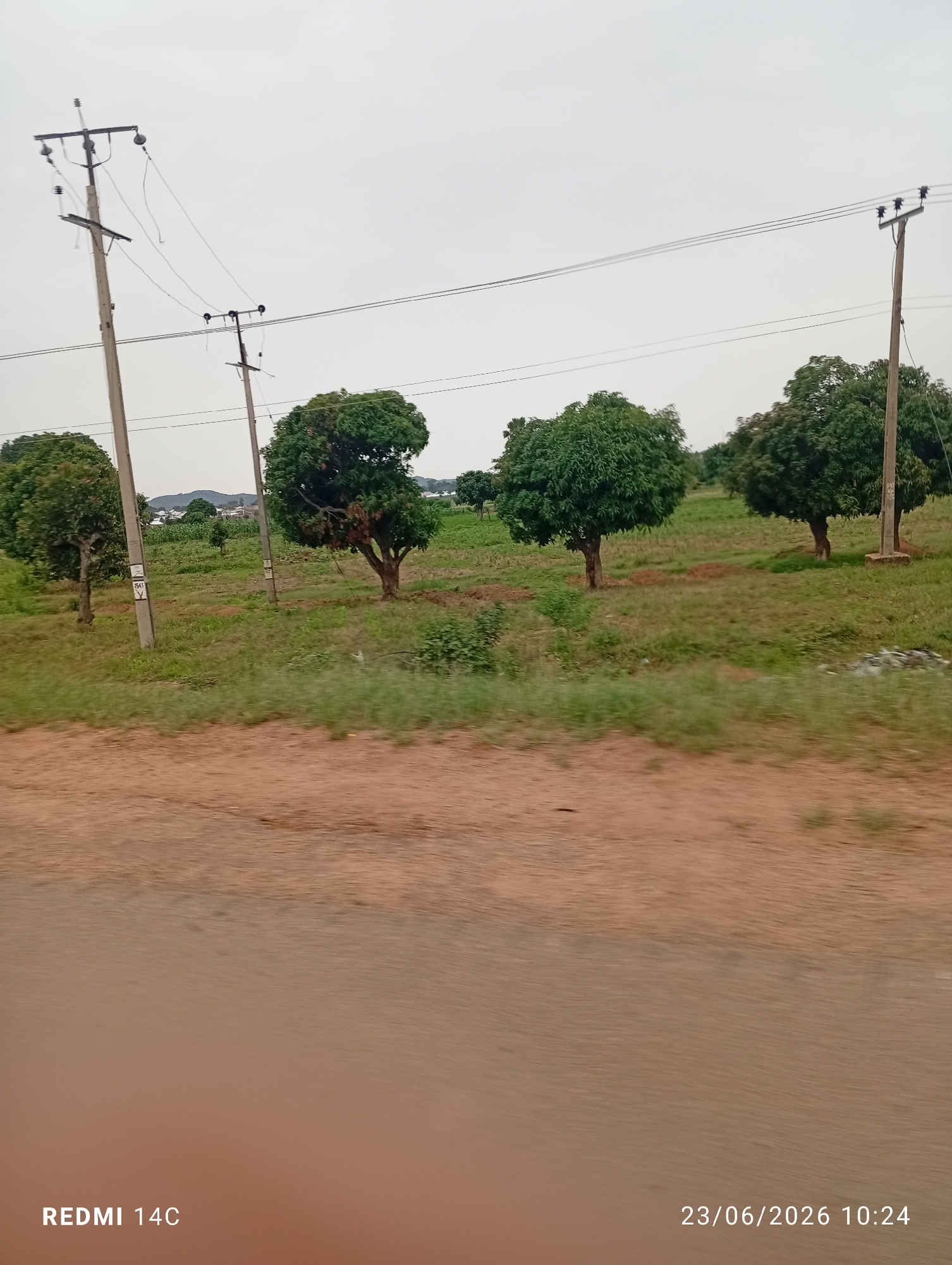 The image depicts an outdoor scene in Du, Nigeria, with several power poles and lines stretching across the frame, indicating an area with electrical infrastructure. In the foreground, a dirt road or path is visible, with a suggestion of movement or travel due to slight blurring. Behind the road, a grassy field with scattered trees occupies the mid-ground. The trees are lush and green, suggesting a temperate or well-watered environment. In the distance, more trees and some buildings can be seen, hinting at a rural or semi-urban setting. The sky is overcast, indicating a cloudy or possibly rainy day, and the time of day appears to be daytime. The overall mood is natural and serene, with no people or specific events clearly depicted. Visible text in the image includes "REDMI 14C" and "23/06/2026 10:24", likely a camera model and timestamp.