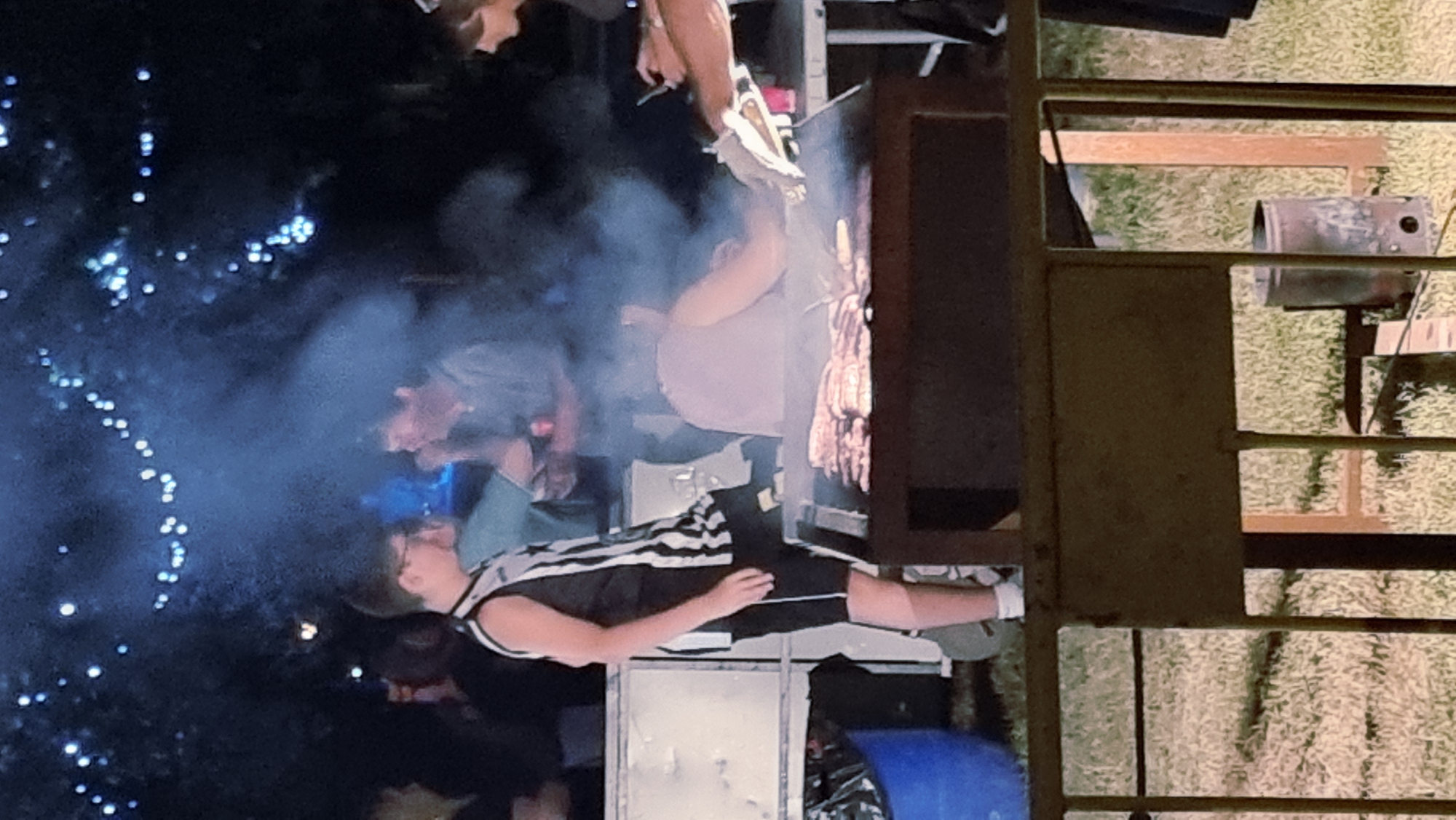 At night, with string lights overhead, a group of people are gathered around a large barbecue grill. The grill is laden with numerous pieces of meat, likely sausages or burgers, cooking in the smoke. A young person in a black and white striped jersey and black shorts stands to the left of the grill. To their right, another individual in a grey shirt is talking to someone out of frame. Behind the grill, a person in a light pink top is tending to the food. To the far right, a person in a dark top is holding what appears to be a phone and a paper towel. The background is dark, suggesting an outdoor setting, with some trees visible. The ground appears to be grass. The scene is likely a public event or gathering in Sant Joan Les Fonts, Spain.