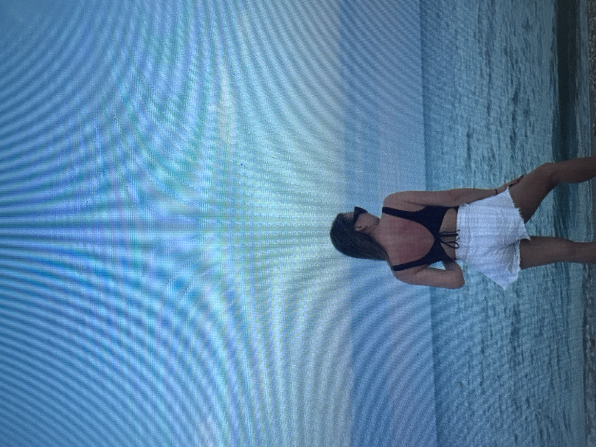 A woman with long brown hair and sunglasses stands with her back to the camera on a beach, facing the ocean. She is wearing a dark bikini top with a crisscross back and white frayed shorts. Her skin appears to be sunburned on her upper back and shoulders. The ocean is a deep blue, with gentle waves. The sky is a pale blue, with some clouds on the horizon. The image appears to have been taken on a digital camera, as evidenced by the screen-like pattern visible throughout the photo. The scene is serene and peaceful, suggesting a relaxing day at the beach.