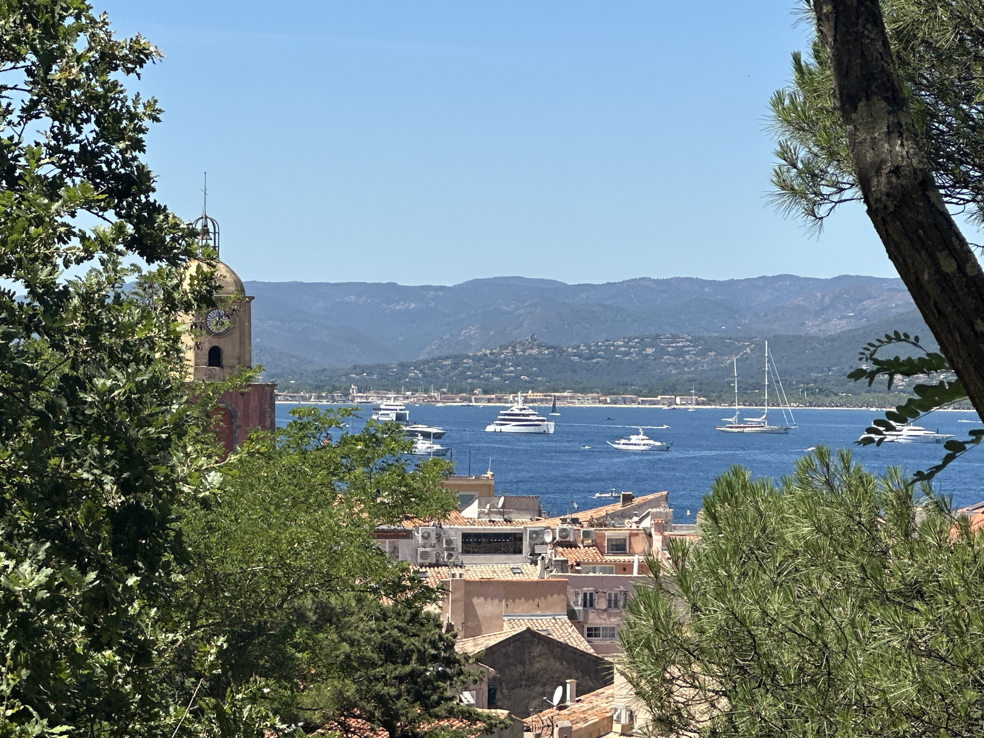 This is a medium shot photograph taken from an elevated position, looking out over the town of Saint-Tropez, France, towards the Mediterranean Sea. The foreground is dominated by lush green foliage from trees, with branches and leaves partially obscuring the view. In the middle ground, the terracotta rooftops of Saint-Tropez are visible, interspersed with buildings showcasing architectural details like air conditioning units and satellite dishes. A prominent bell tower with a clock face, part of a church structure, stands to the left of the frame. The background features the deep blue of the sea, dotted with numerous yachts and sailboats of varying sizes. Beyond the sea, a range of hazy blue mountains forms the horizon line under a clear, bright sky. The scene appears to be set on a sunny day, evoking a coastal, Mediterranean atmosphere. There are no discernible people in the image.