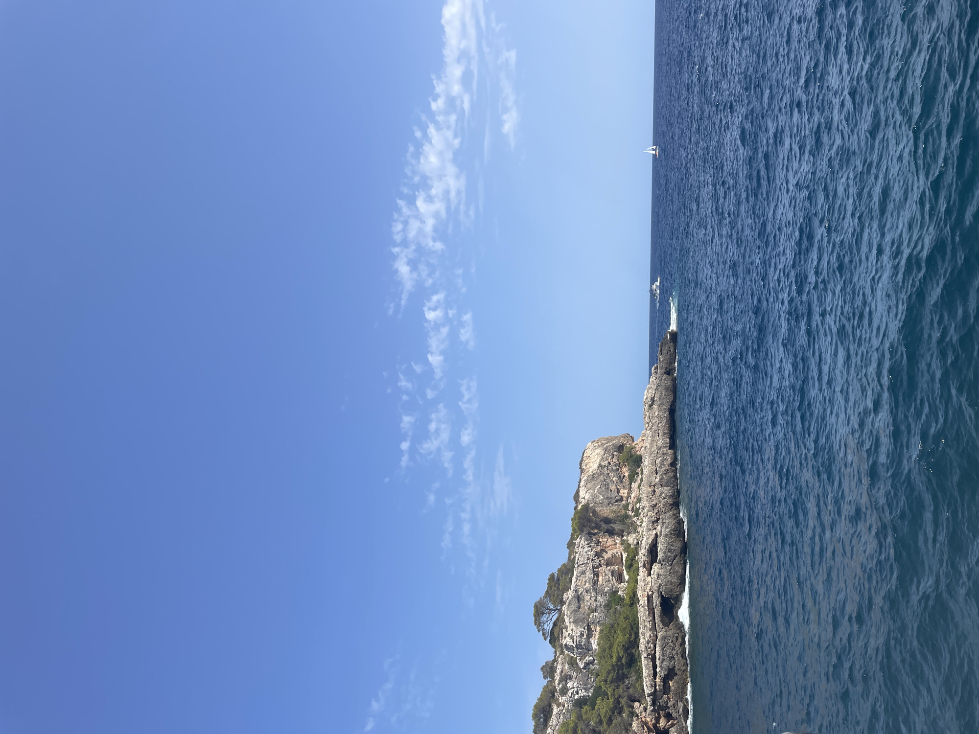 A wide, horizontal shot captures a bright, sunny day at sea off the coast of Santanyí, Spain. The deep blue ocean occupies the lower half of the frame, with gentle ripples on its surface. In the distance, a rocky, tree-covered cliff juts out into the water on the left. A lone sailboat with a white sail is visible on the right side of the image, and a smaller boat is nearer to the cliffs on the left. The upper half of the image is dominated by a clear, bright blue sky, with a scattering of wispy white clouds. The scene evokes a sense of peace and natural beauty.