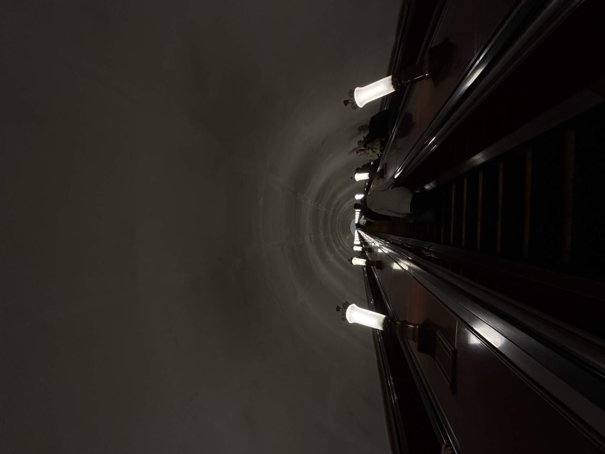 The image shows the interior of a Moscow Metro station. An escalator descends into a cylindrical tunnel, illuminated by a series of evenly spaced, vintage-style lamps along the tunnel wall. Several people are riding the escalator. The walls are plain and appear concrete, while the escalator's railings and fixtures have a metallic sheen. The overall atmosphere is dimly lit.