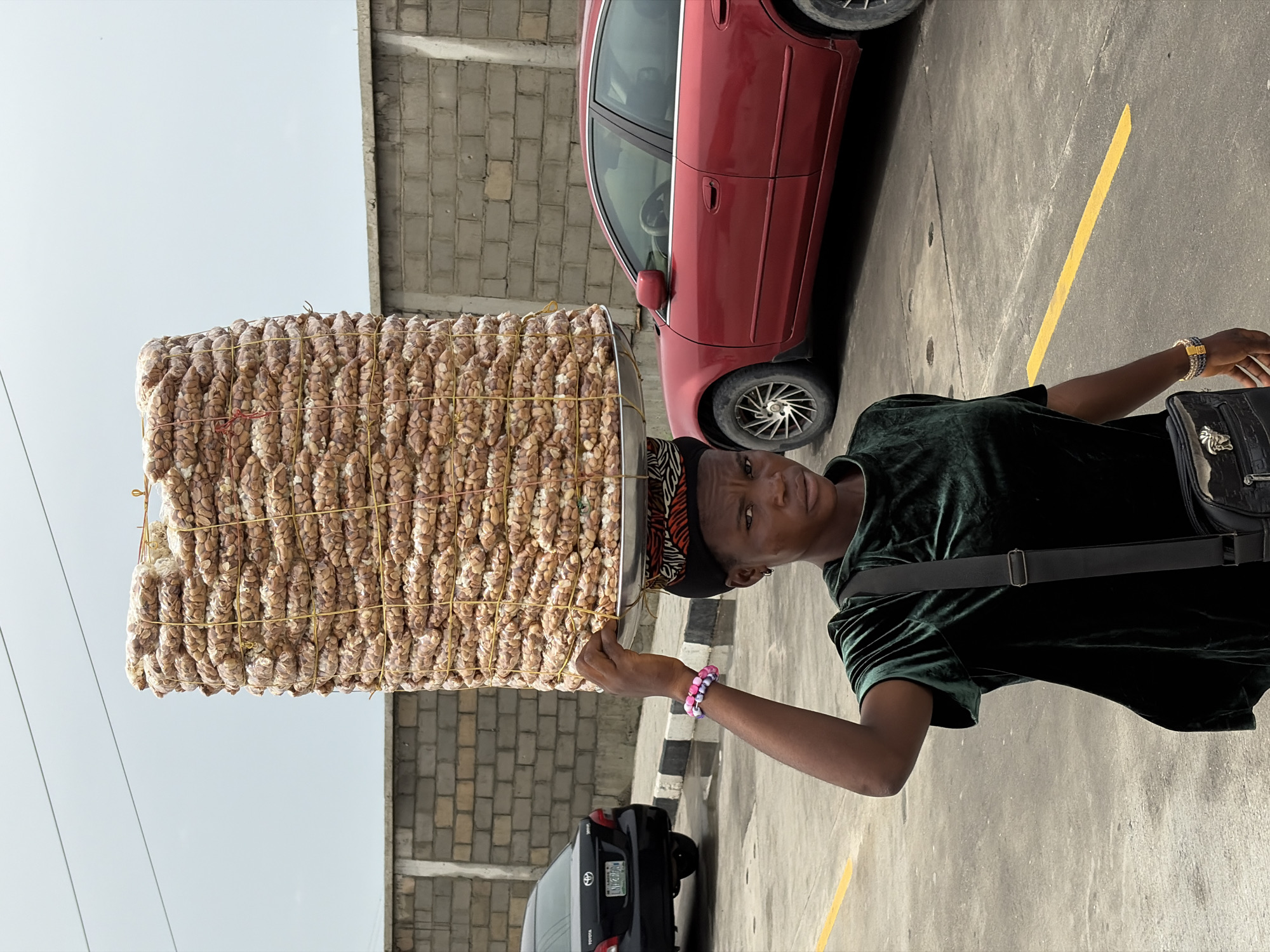 A street vendor in Igboefon, Nigeria, is balancing a large tray stacked high with small packages, likely of food or snacks, on their head. The packages are arranged vertically and secured with yellow twine. The vendor is wearing a dark green velvet shirt, a patterned head covering, and bracelets on their left wrist. They also have a black shoulder bag. The background includes a red car, a black car, a grey brick wall, and a concrete pavement with a yellow line marking. The scene appears to be an outdoor parking area or street.