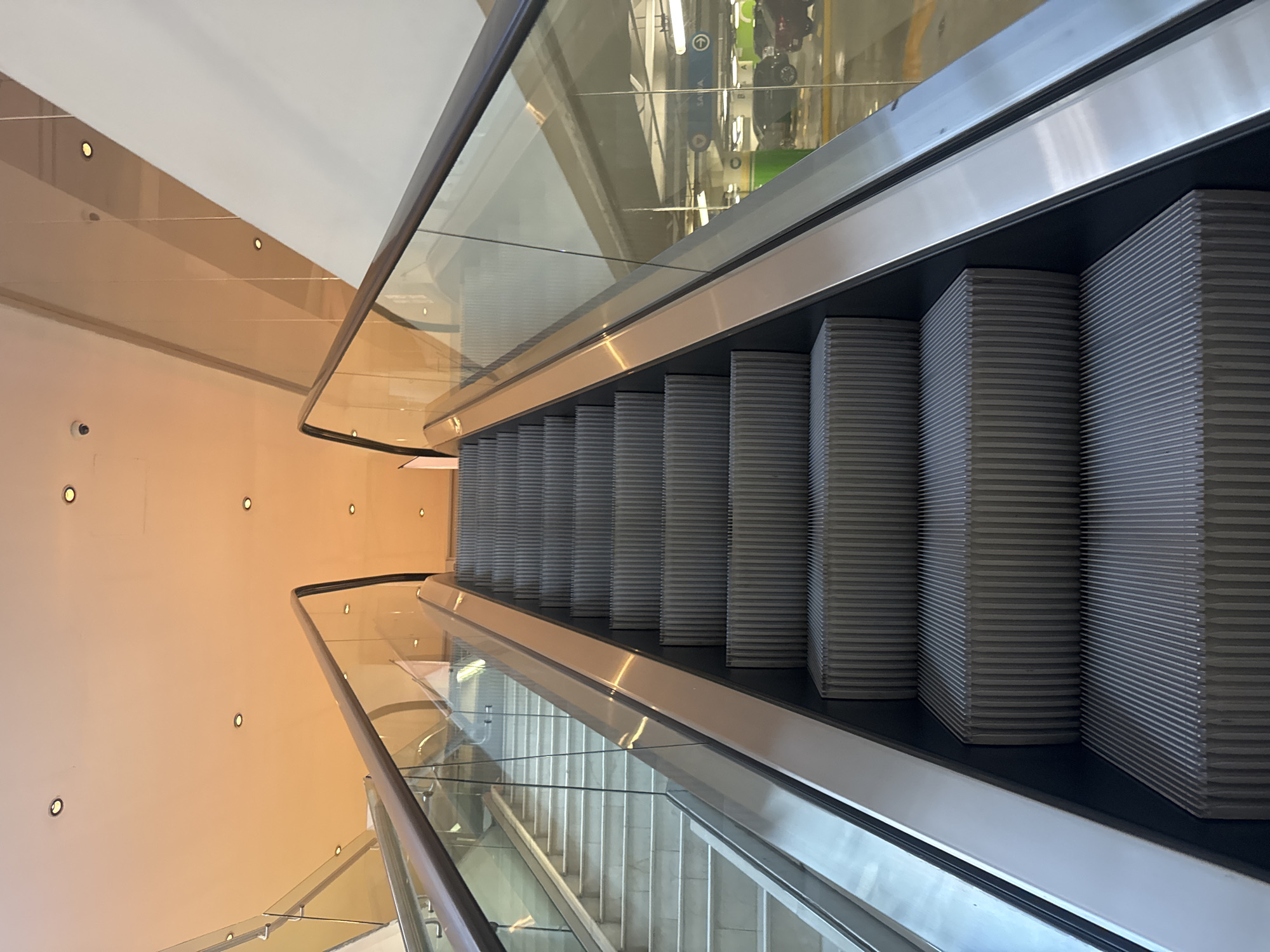 The image depicts an escalator descending between floors inside a building. The escalator has gray steps and glass sides with metal handrails. The adjacent walls are peach-colored with recessed lighting. Reflections in the glass sides show the floor above, with glimpses of green and yellow objects. The location is Puebla, Mexico. No people are visible in the image.