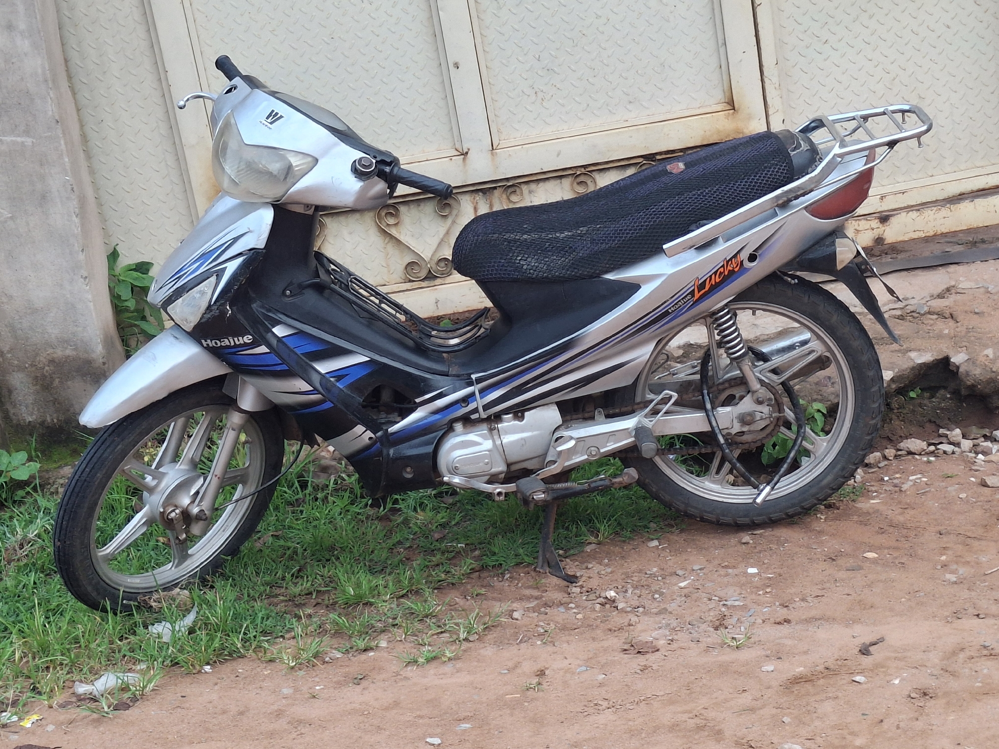 A silver and black motorcycle is parked on a dirt and grass area in front of a metal gate. The motorcycle has a "Lucky" decal on its side. The front wheel is on the left and the rear wheel is on the right. The motorcycle is upright, supported by its kickstand. The background consists of a textured metal gate and some foliage. The ground is composed of reddish-brown dirt and patches of green grass. The scene appears to be outdoors, possibly in an urban or semi-urban setting.