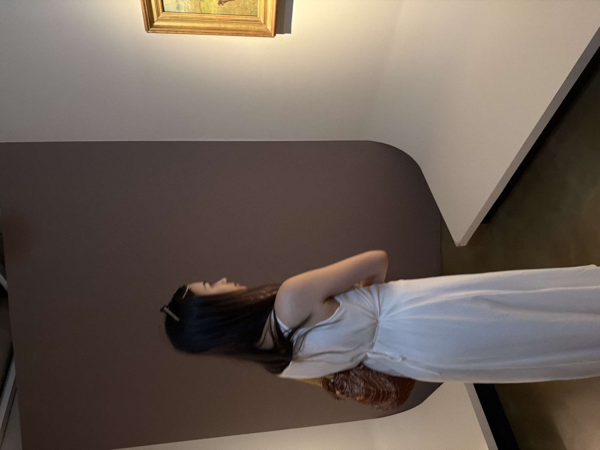 A woman in a white dress stands in an art gallery, looking at a framed painting. The gallery has a brown wall behind the woman and a cream-colored curved wall to her right. The painting is on the cream-colored wall, and it appears to be a landscape with a gold frame. The woman is holding a woven bag on her left shoulder. Her hair is dark and pulled back. She is wearing sunglasses on top of her head. The lighting in the gallery is dim. There are no other people visible in the image. The location is not specified in Seoul, South Korea.