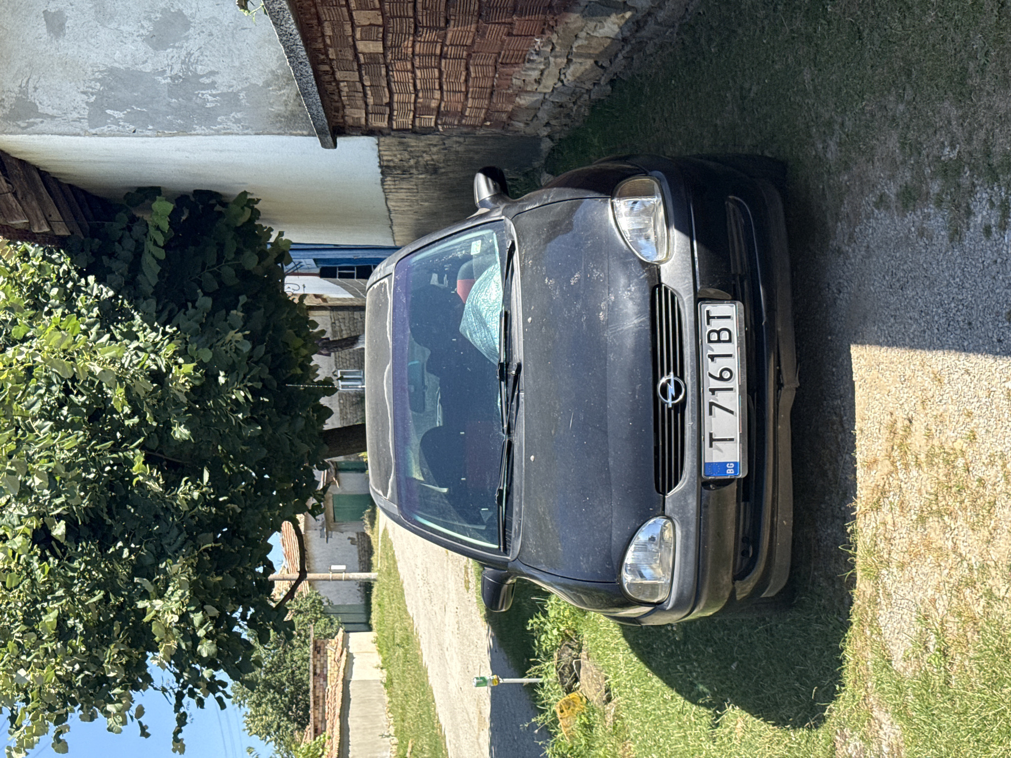 A black Opel Corsa is parked on a gravel driveway in front of a building with a brick wall. The car is facing forward, and its license plate reads "T 7161 BT" with "BG" indicating Bulgaria. A large tree with green leaves partially overhangs the car and the building. The surrounding environment appears to be a rural or village setting, with a dirt road leading away from the car and other buildings visible in the background. The lighting suggests it is a sunny day.