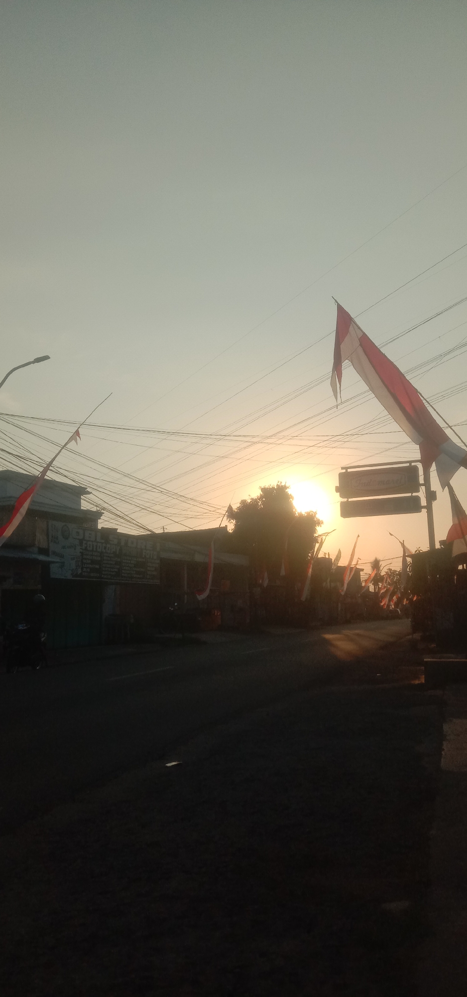 The image depicts a street scene at sunrise in Cangkringrandu, Indonesia. The sky is a pale orange and yellow, indicating the early morning sun. Red and white Indonesian flags are strung across the street and are attached to poles along the side of the road. Several power lines crisscross the scene. In the foreground, the road is dark and appears to be made of asphalt. On the left side of the street, there are buildings with signs, including one that reads "OBL STORE I" and "FOTOCOPY". A motorcycle is parked in front of one of the buildings. On the right side of the street, there is a sign that reads "Indomaret". Trees are visible in the background, silhouetted against the bright sun. The overall impression is a quiet, rural or suburban street preparing for the day.