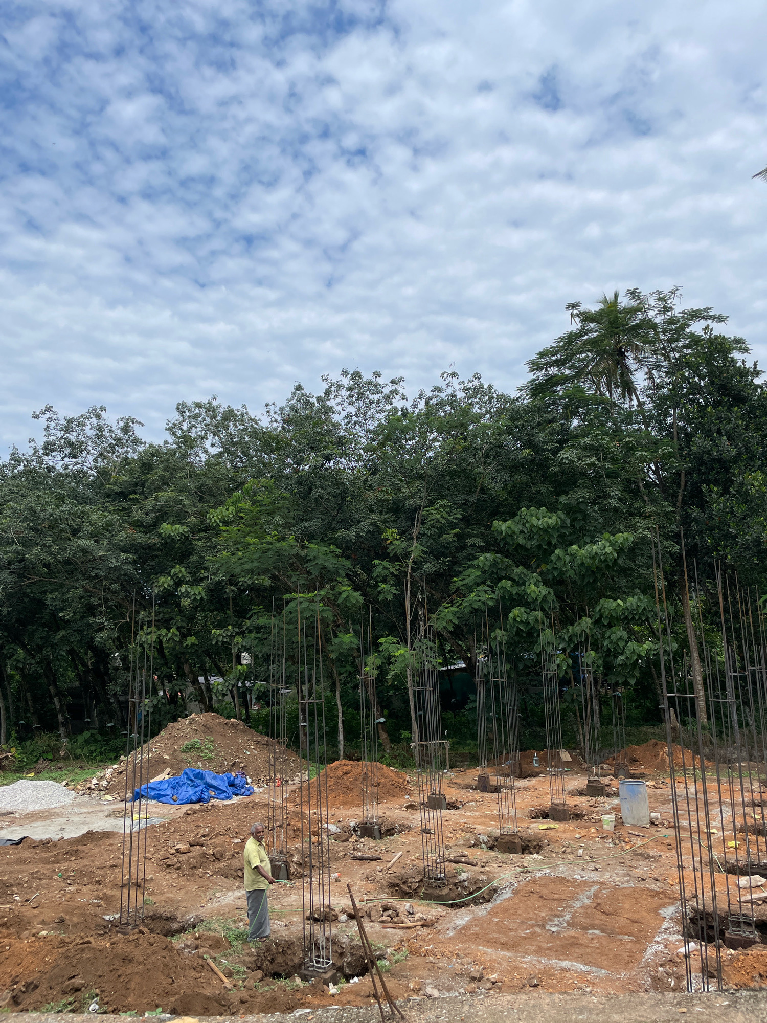 The image depicts a construction site in Kudappanamoodu, India. The foreground shows the foundation work for a building, with numerous rebar pillars emerging from the earth. The ground is uneven, composed of red soil and scattered rocks. A man in a light yellow shirt and grey pants is visible in the center-left, holding a green hose, possibly watering the area. Blue tarps are spread over a pile of materials in the middle ground. In the background, dense green foliage, including trees and palm trees, forms a natural barrier. The sky above is partly cloudy with scattered white clouds against a light blue backdrop.