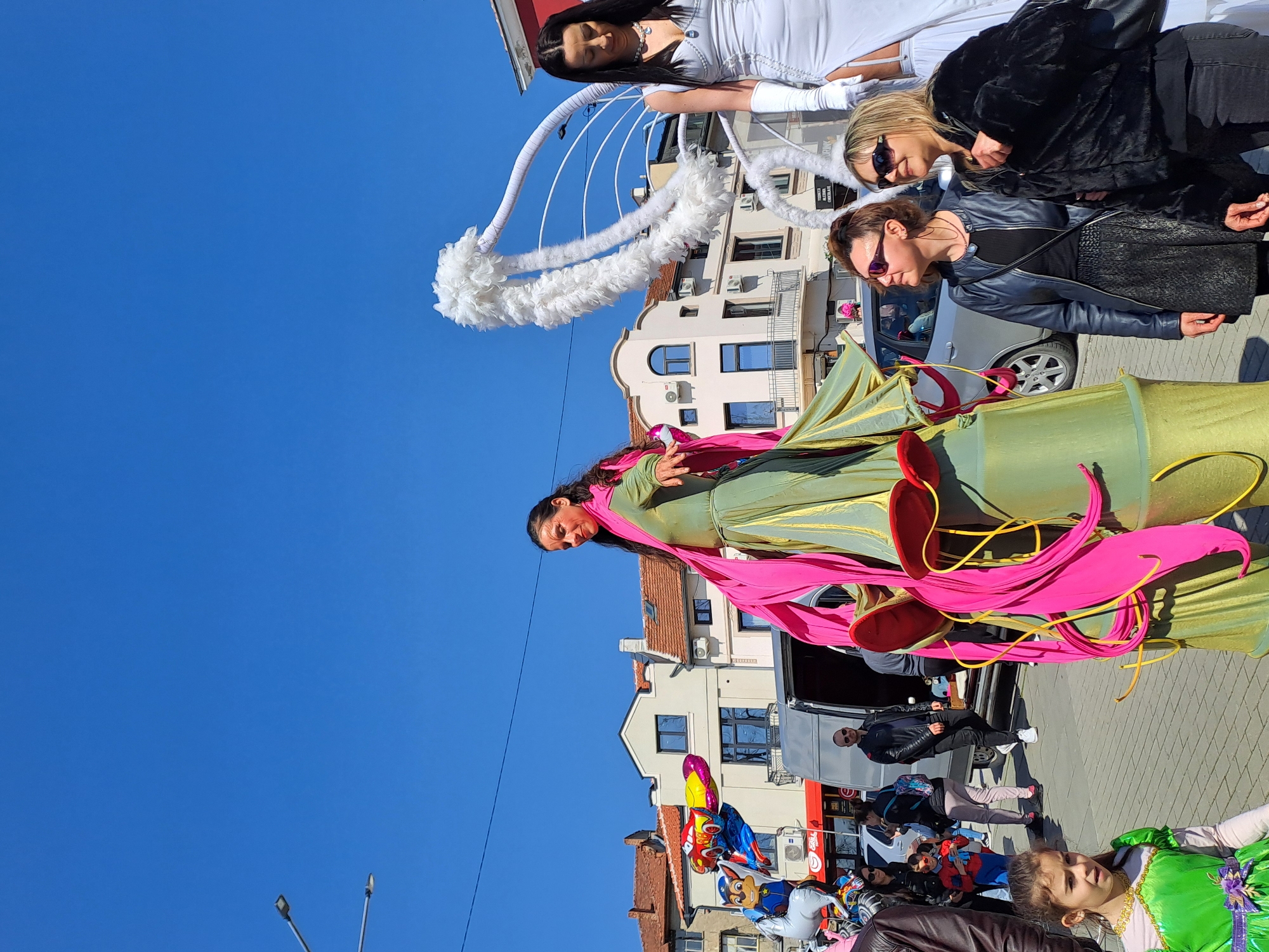 The image shows a street scene in Veliko Tarnovo, Bulgaria. The sky is clear and blue. In the foreground, a woman stands tall, elevated, wearing a long green dress with pink ribbons and adornments. Next to her are other women and children, some dressed in costumes. There are buildings, street lights, and overhead power lines visible in the background. Balloons and other celebratory decorations are apparent along the street. Cars are parked along the street and some people sit or walk near a store with a red awning. The overall scene suggests a street celebration or parade.