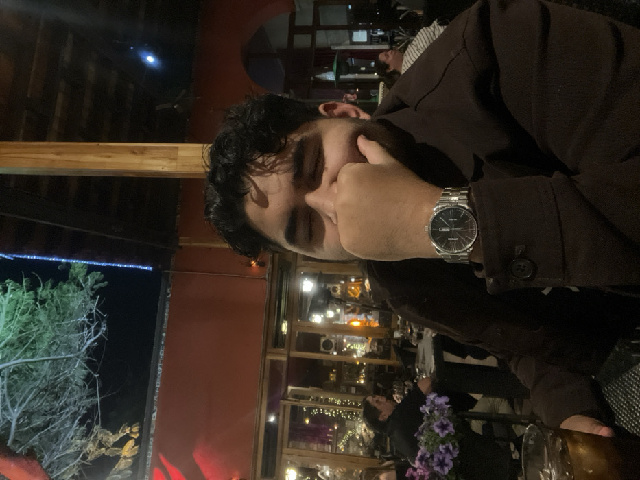 A man with dark curly hair and a brown jacket is seated at a restaurant at night. He is looking at his silver wristwatch and resting his face on his hand. In the background, blurred figures are visible, along with a window reflecting the interior lights. A tree with string lights is partially visible to the left.