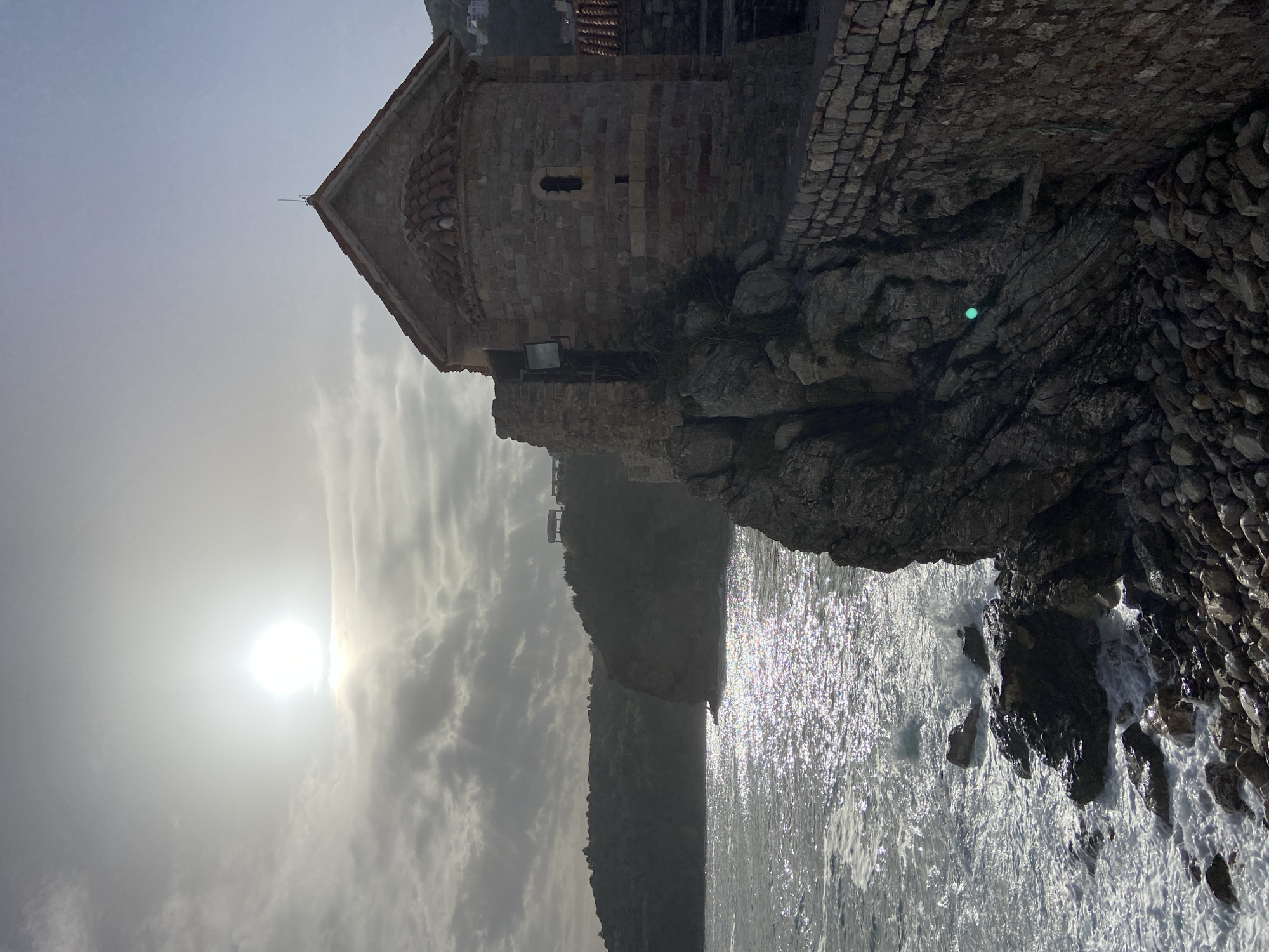 The image shows a stone building with a red-tiled roof, possibly a church or chapel, situated on rocky terrain next to the sea. The sun is positioned in the upper left portion of the frame, creating a hazy, bright sky. The rocky cliff extends into the water where waves are crashing. A hazy mountain is visible in the middle ground, extending into the sea. The building is built into the cliffside and constructed of grey stone. The location is Sveti Stefan in Budva, Montenegro.