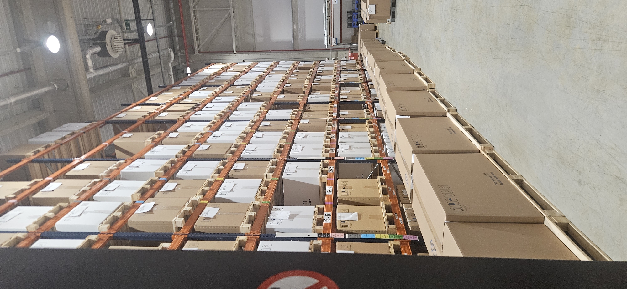 The image depicts a warehouse interior, possibly located in Plovdiv, Bulgaria, given the directive. Multiple rows of shelving units filled with cardboard boxes are visible. The boxes appear to be uniformly stacked on pallets. The warehouse has a high ceiling with visible lighting fixtures and ventilation systems. The floor is concrete. The image is taken from a slightly elevated perspective. At the bottom of the image, a portion of an object, possibly a barrier, is visible, featuring a no-entry symbol.