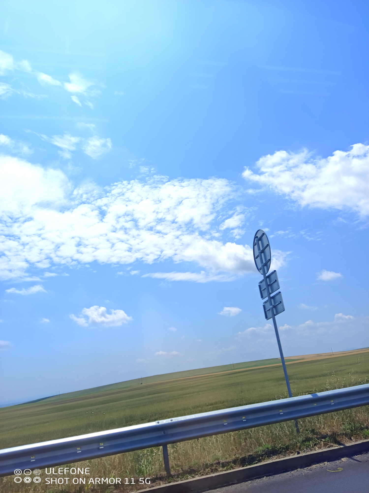 The image depicts an outdoor daytime scene captured on a sunny day with a clear blue sky and scattered white cumulus clouds. A prominent lens flare is visible in the upper left quadrant of the sky.
The landscape consists of gently rolling hills covered predominantly in green agricultural fields, with some lighter yellowish-brown sections indicating different crops or drier areas. In the far distance on the hills, several thin, vertical poles, possibly utility poles, are faintly visible against the horizon.
In the mid-ground, a metallic road sign assembly is positioned to the right of the frame. It consists of a circular sign at the top and two rectangular signs stacked vertically below it, all mounted on a single grey metallic pole. The signs are viewed from the back and appear dark grey or translucent blue, revealing structural details on the circular sign.
The foreground is dominated by a silver-colored metallic guardrail that extends horizontally across the lower third of the image, angling slightly upwards from left to right. The guardrail is composed of multiple segments bolted together. Below the guardrail, a narrow strip of roadside vegetation, primarily dry grasses and weeds, is visible. The edge of a dark asphalt road surface is present in the lower right, bordered by a concrete curb which has a faint yellow mark on its surface.
The bottom left corner of the image contains a watermark with the text "ULEFONE" and "SHOT ON ARMOR 11 5G", accompanied by several circular icons.
The scene is devoid of any visible people, animals, or active interactions. The location is Unknown, Unknown.